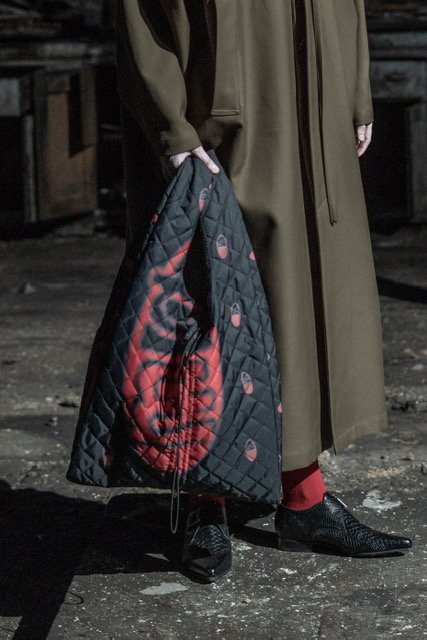Person standing outdoors at night, wearing a long brown coat, red pants, and black shoes, holding a black quilted bag with red graffiti-style artwork.