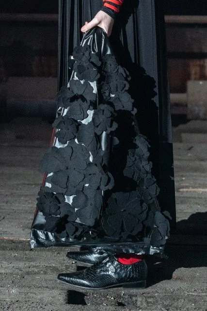 Person holding a black handbag with large black flower appliques, wearing black pointed shoes with red accents, standing on a wooden surface.