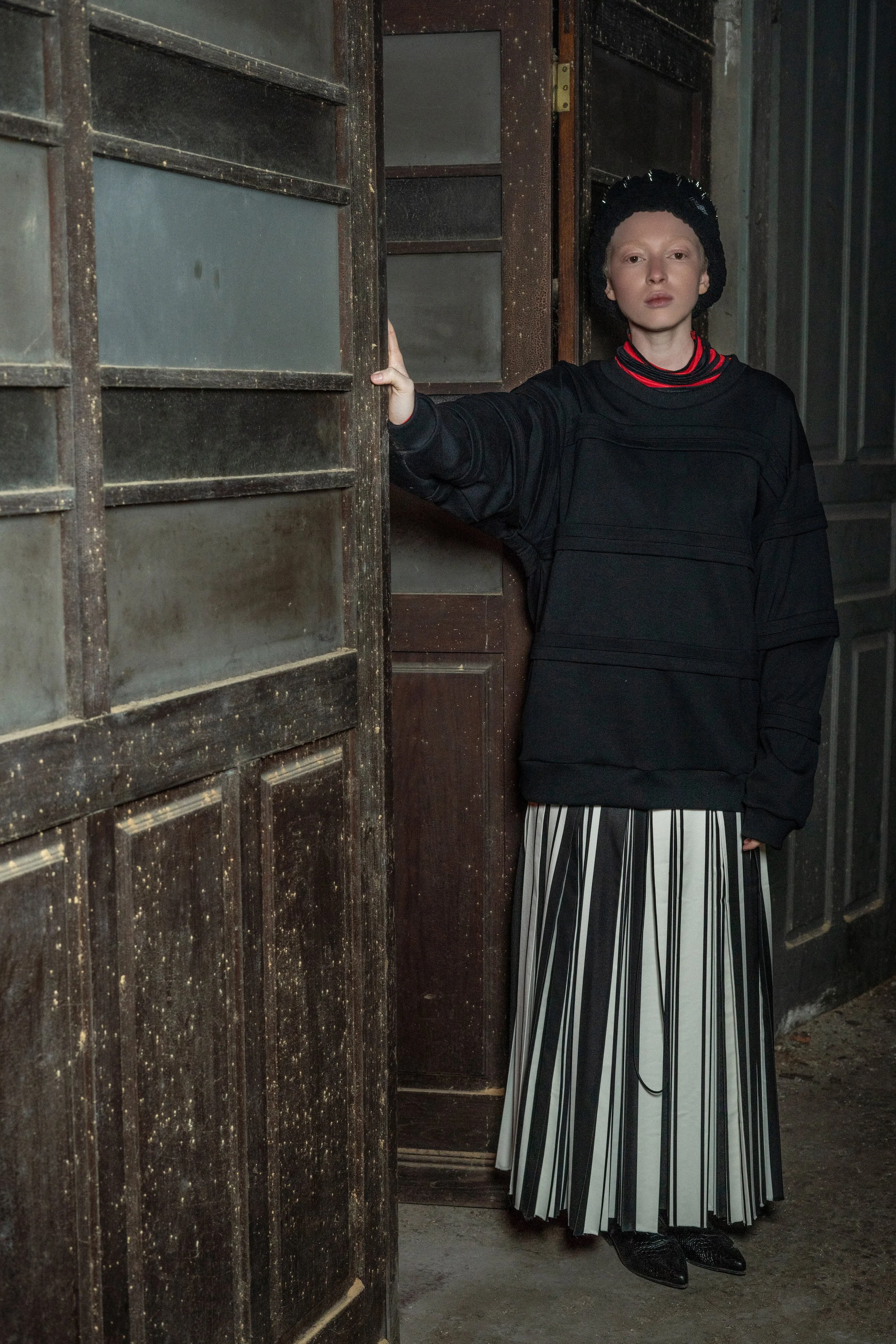 A person with pale skin, short light hair, and a serious expression, standing inside an old, rustic wooden building, wearing a black sweater, black beanie, striped skirt, and black shoes, with one hand touching a weathered wooden door.