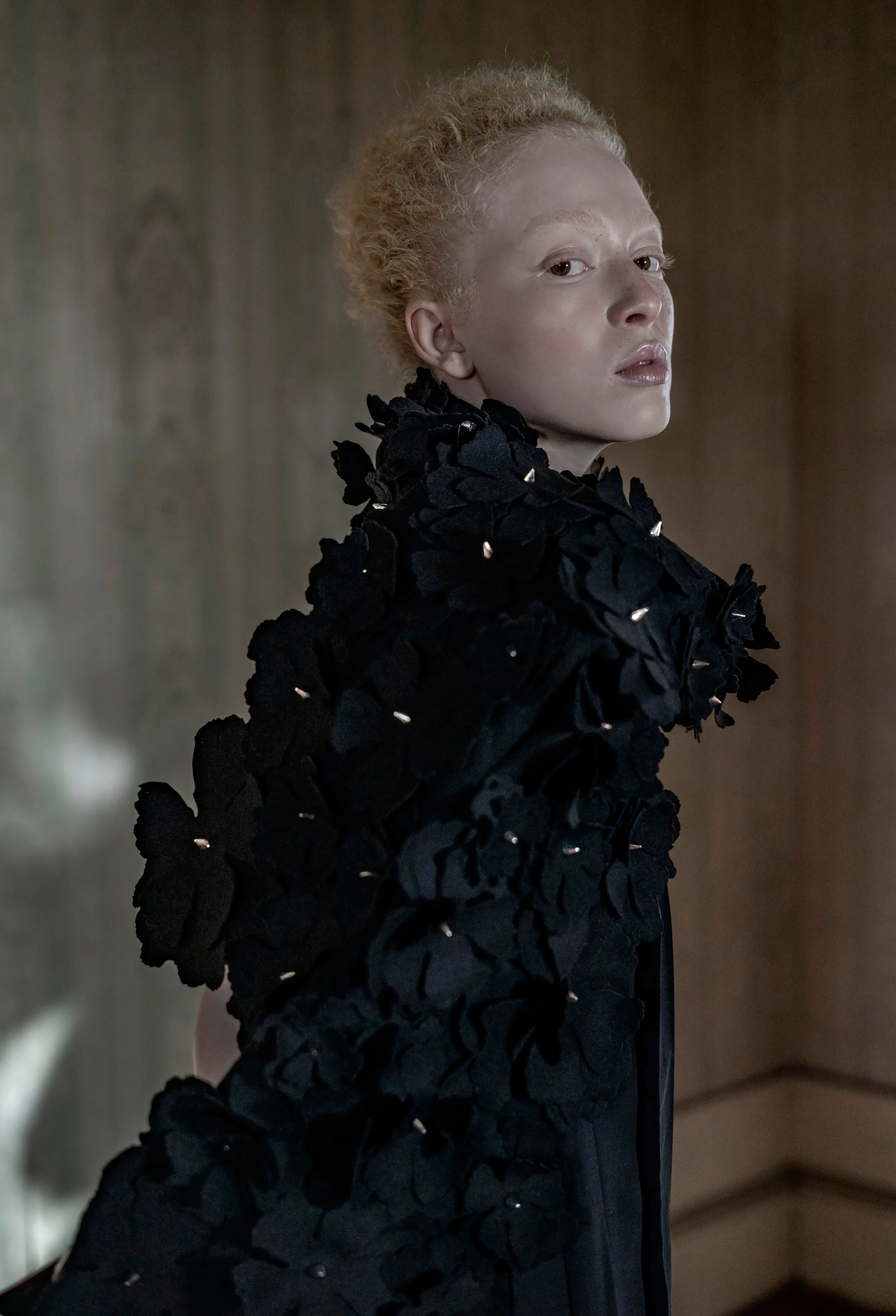 A woman with light skin and curly blonde hair wearing a black textured garment with floral embellishments.