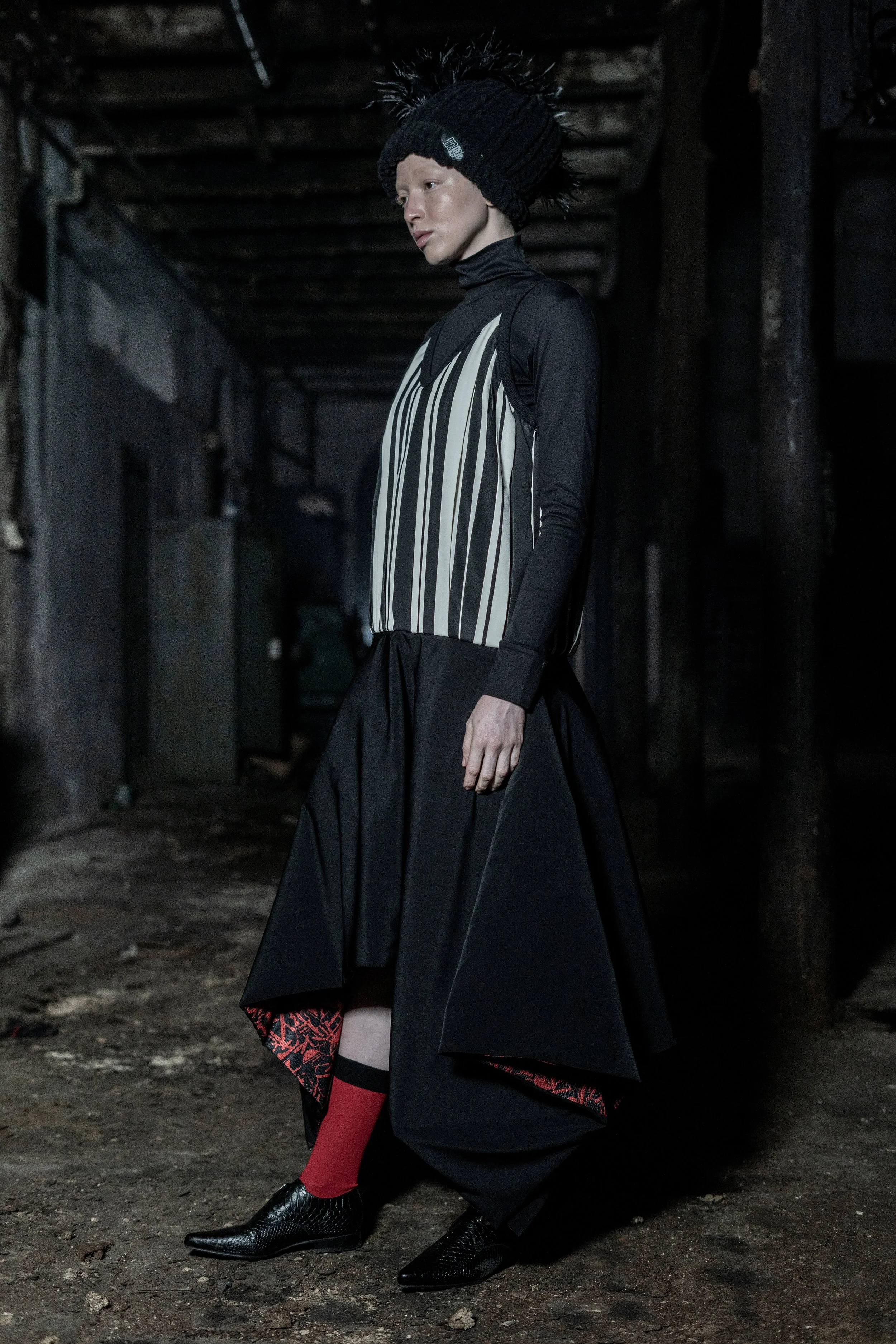 A person wearing a black, shiny textured hat with pom-poms, a black turtleneck, a striped sleeveless top, a black skirt, red socks, and black textured shoes, standing in a dimly lit industrial or warehouse setting.