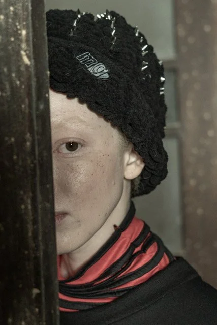 A person with fair skin, light-colored eyes, and freckles wearing a black knit hat and a red and black striped neck gaiter, partially obscured by a wooden surface, with a background of curtains and a wall.