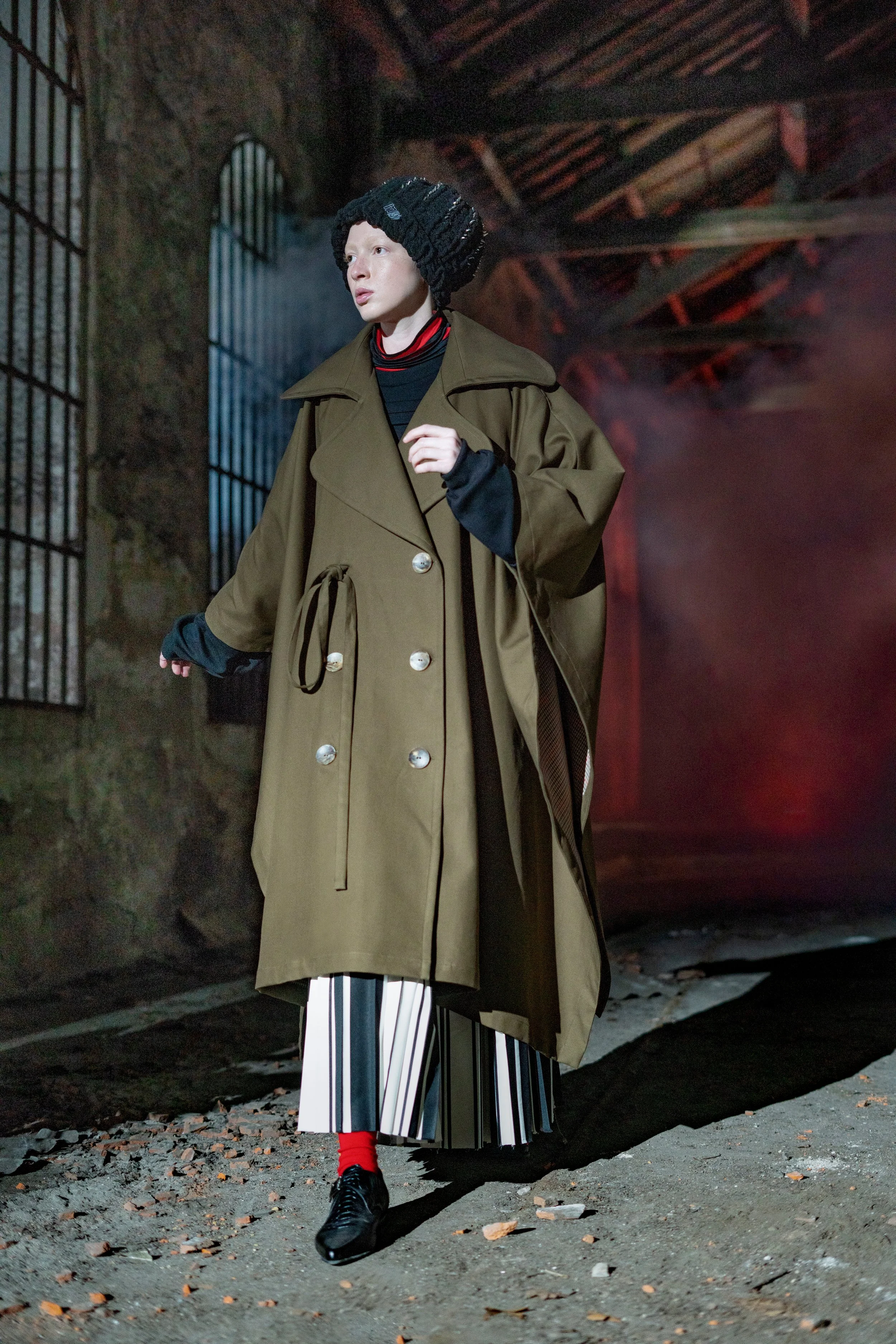 Person in a long brown trench coat, striped skirt, black shoes, red socks, and knit hat, walking outdoors at night.
