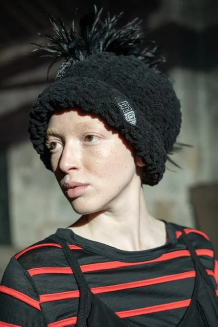 Woman wearing a black, fluffy beanie with a spiked feather decoration and a striped black and red shirt.