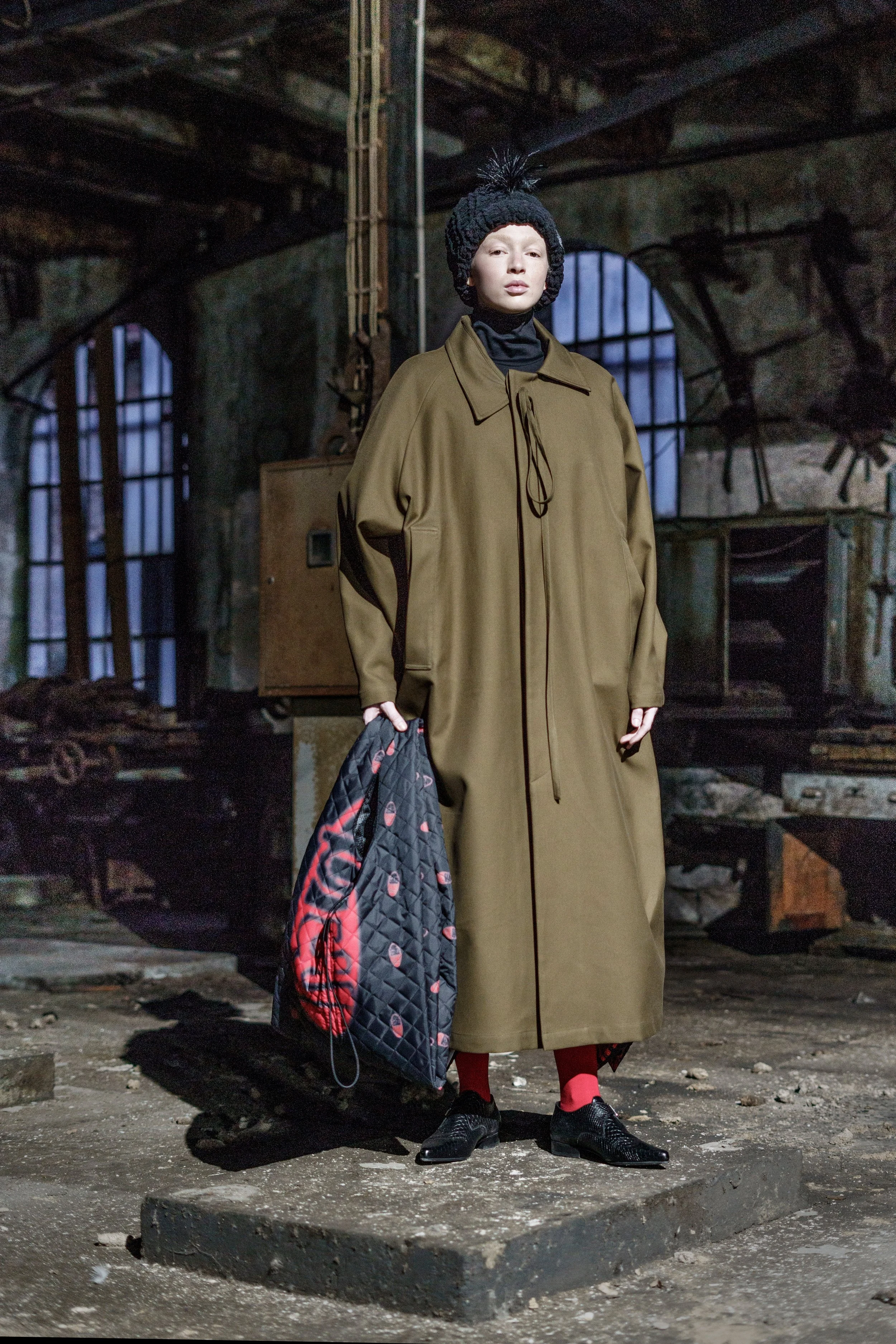 A person standing in an industrial, abandoned building, wearing a long brown coat, black shoes, red socks, a black knit hat with a pom-pom, and holding a black bag with red and gray design.