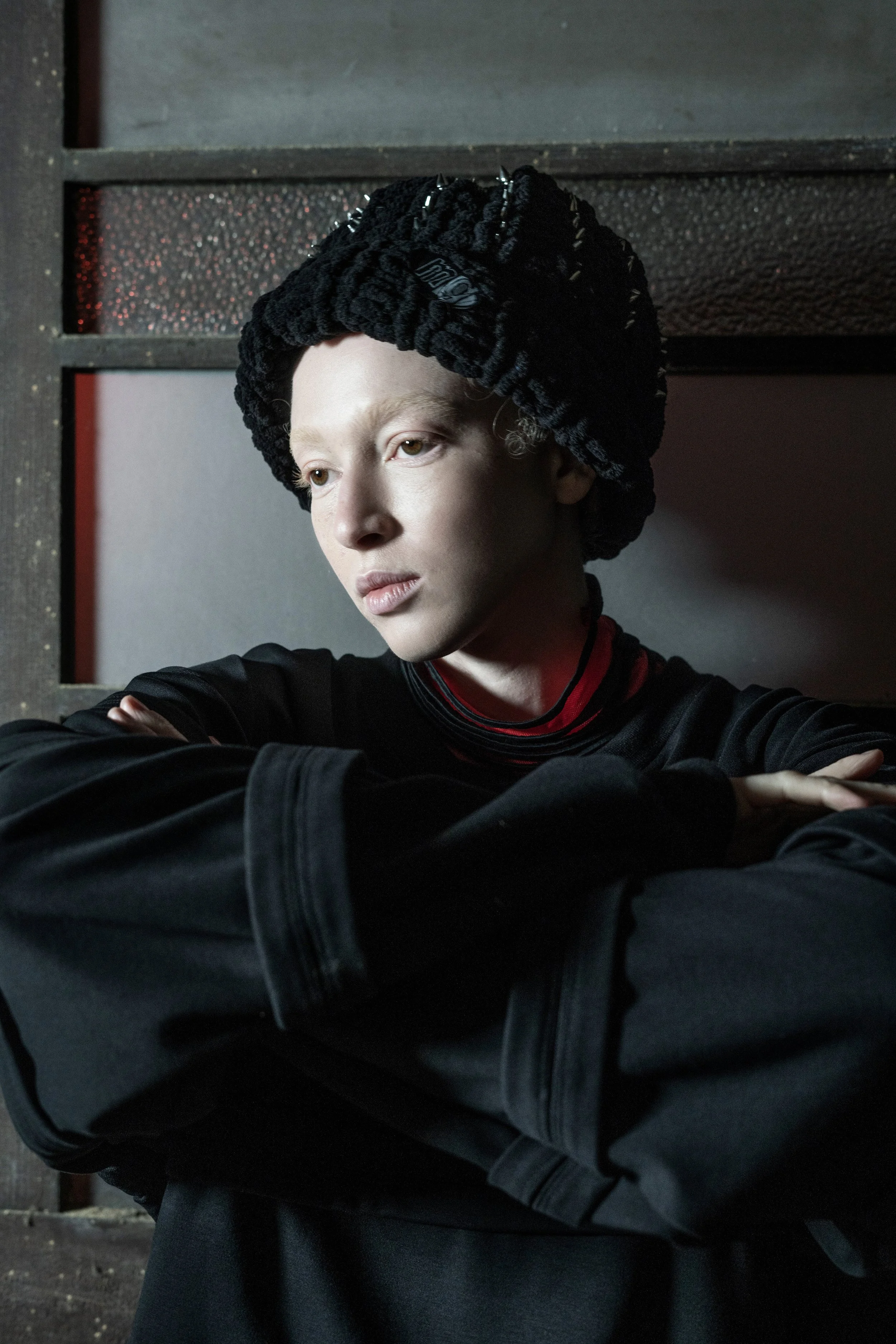 A woman with pale skin, blonde hair, and a, black knit hat looks off to the side with a contemplative expression, arms crossed, wearing dark clothing, background with mixed dark textures.