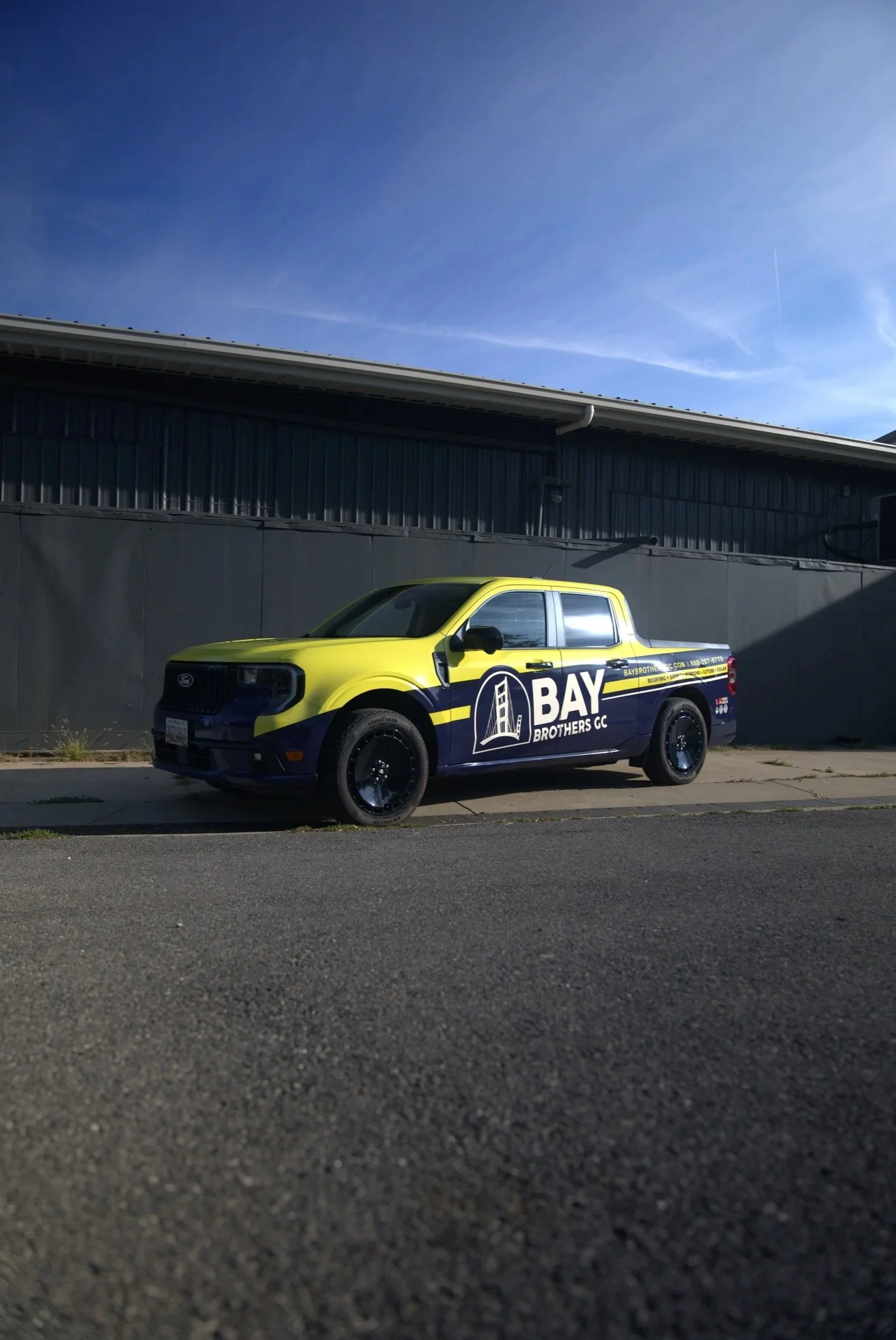 Yellow and black pickup truck with BAY Brothers CC logo parked on pavement in front of a black building, under a blue sky.
