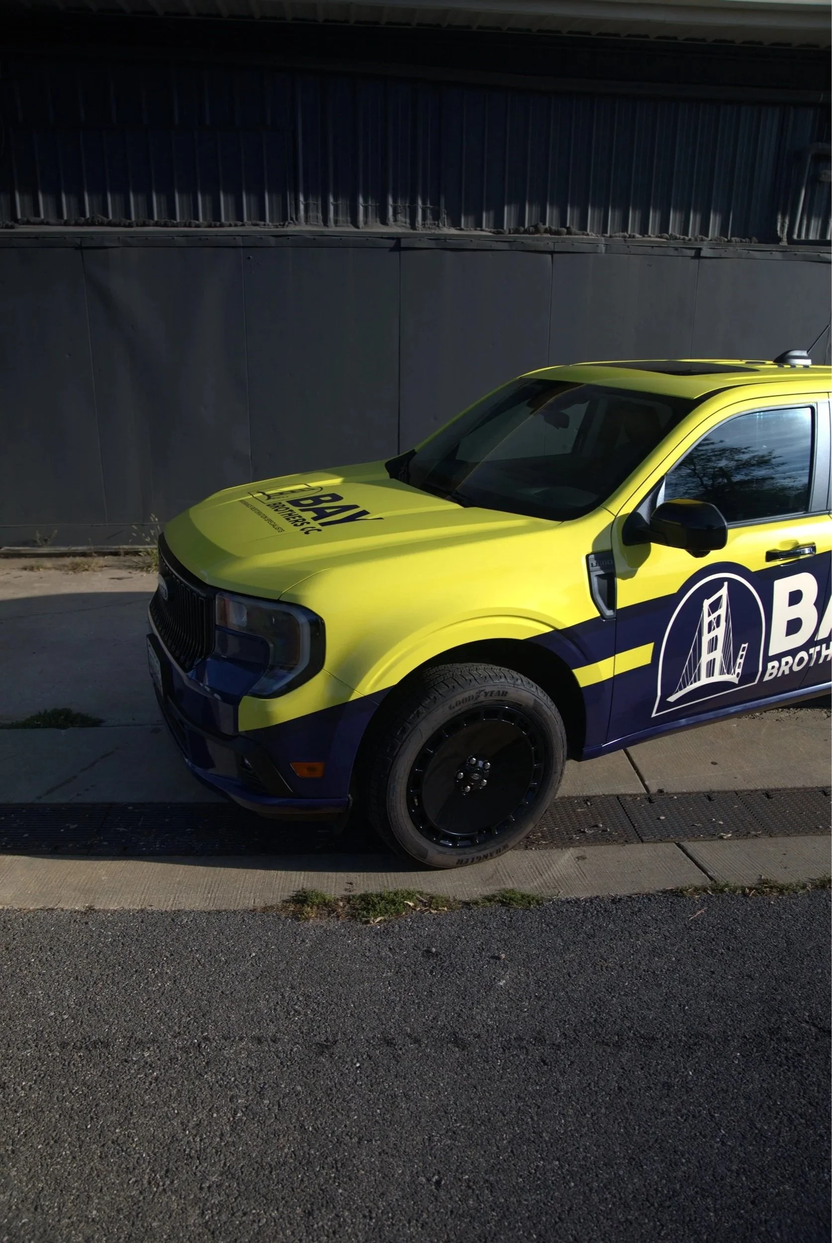 A yellow and black vehicle with a logo of a bridge and the words 'Brothers' on the side, parked on a sidewalk in front of a black metal fence.