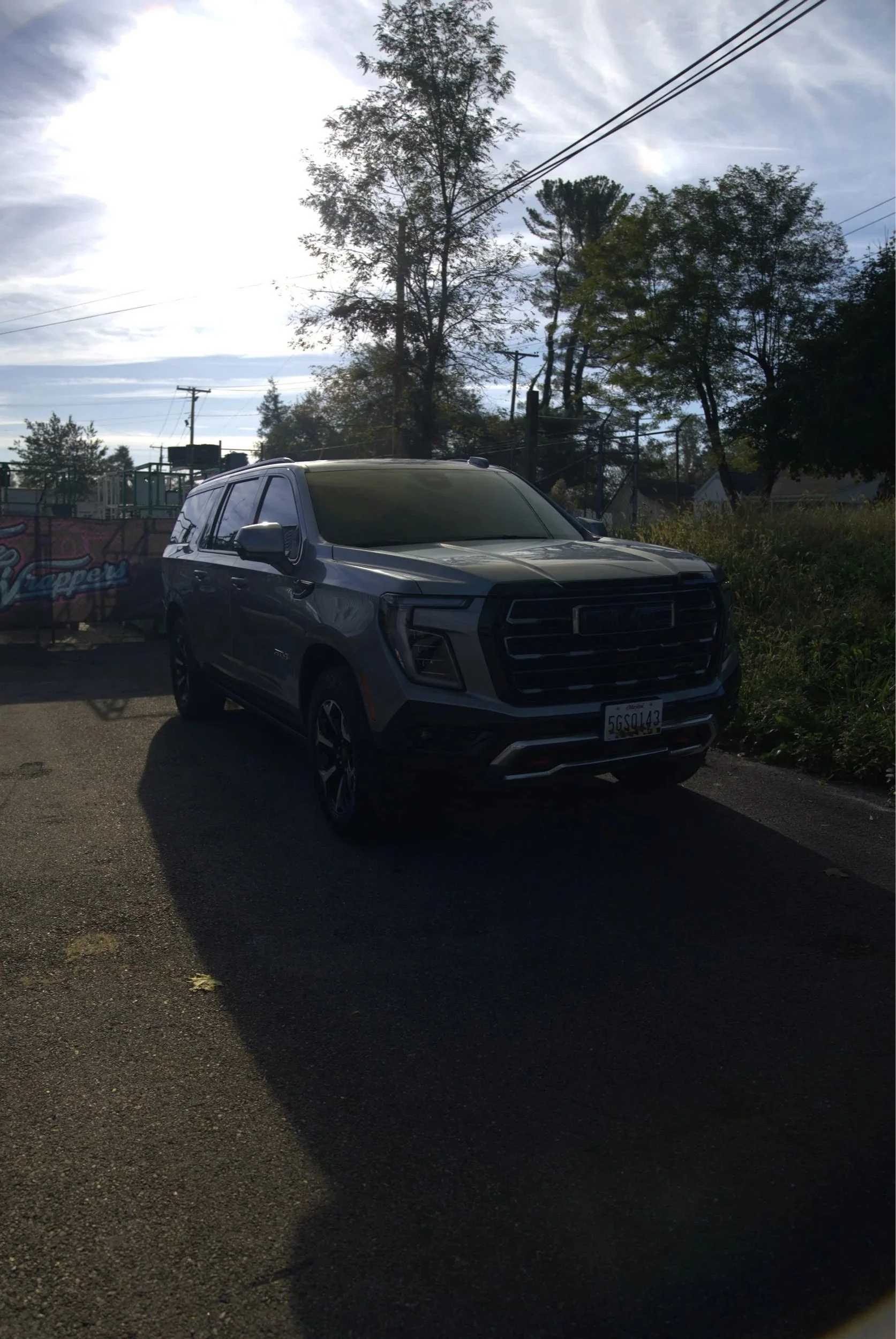 A dark gray or black SUV parked on a paved parking lot with trees and power lines in the background, and the sun shining brightly, creating a shadow of the vehicle on the ground.