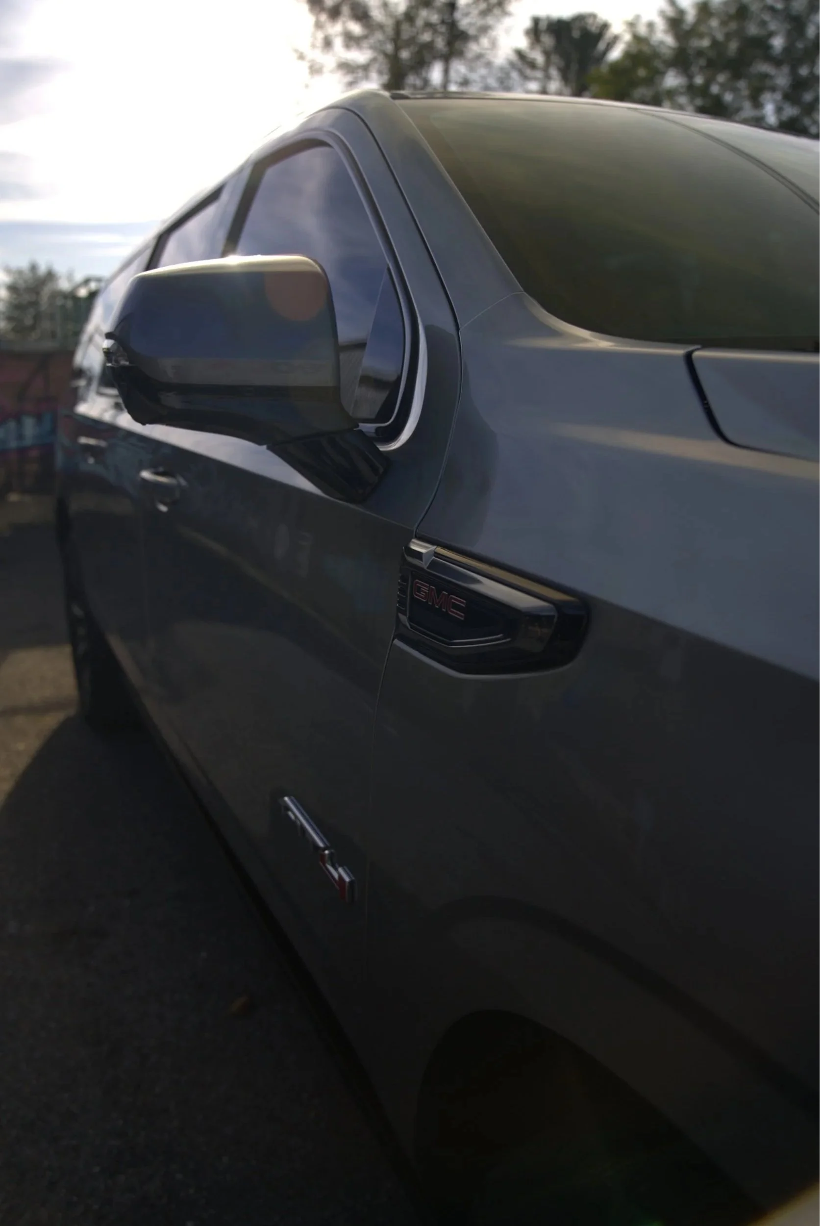 Close-up of a gray GMC vehicle's side, showing part of the front door, side mirror, and GMC badge. The background has trees and a partly cloudy sky.