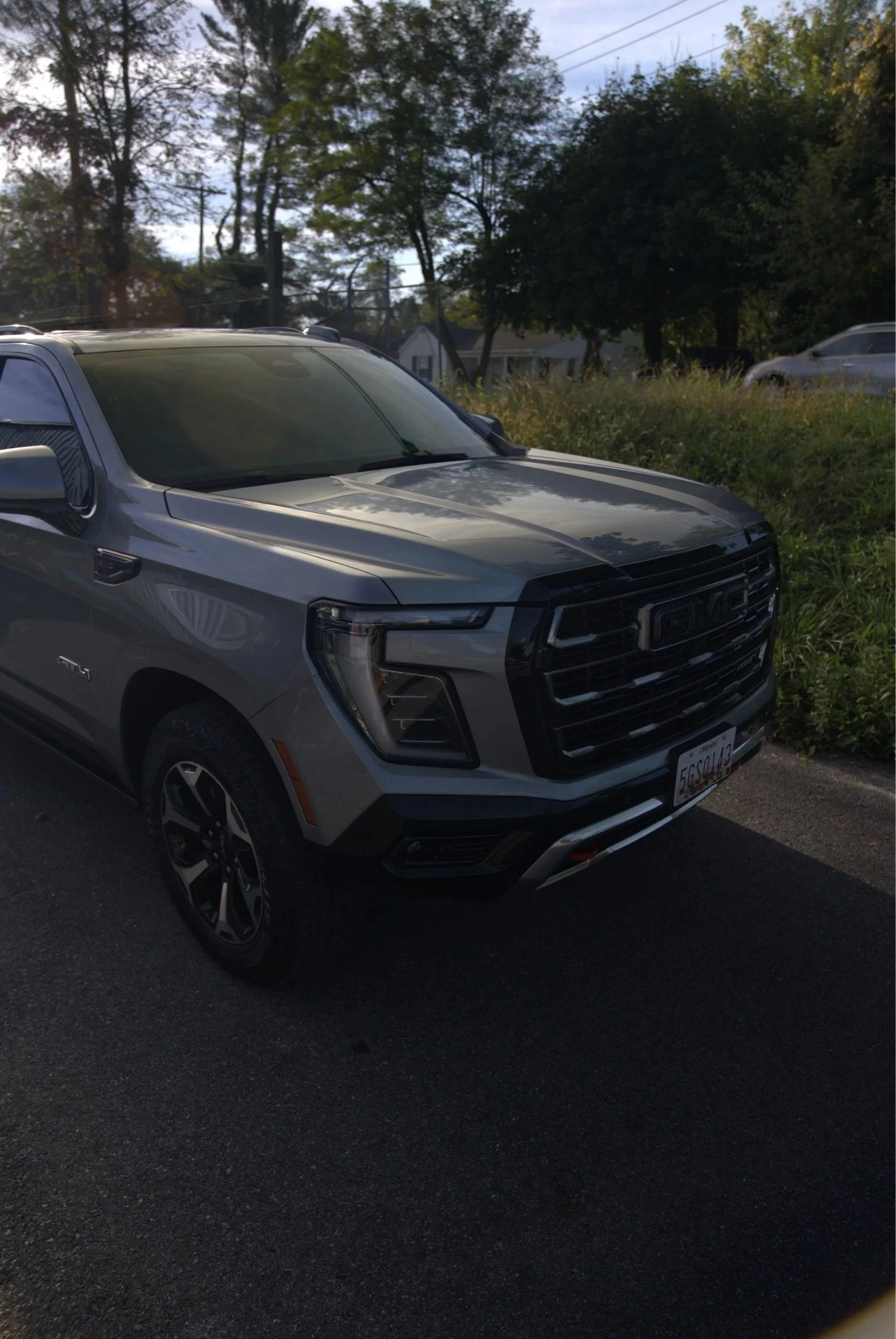 A silver GMC SUV parked on a street with trees and houses in the background, and other vehicles visible.