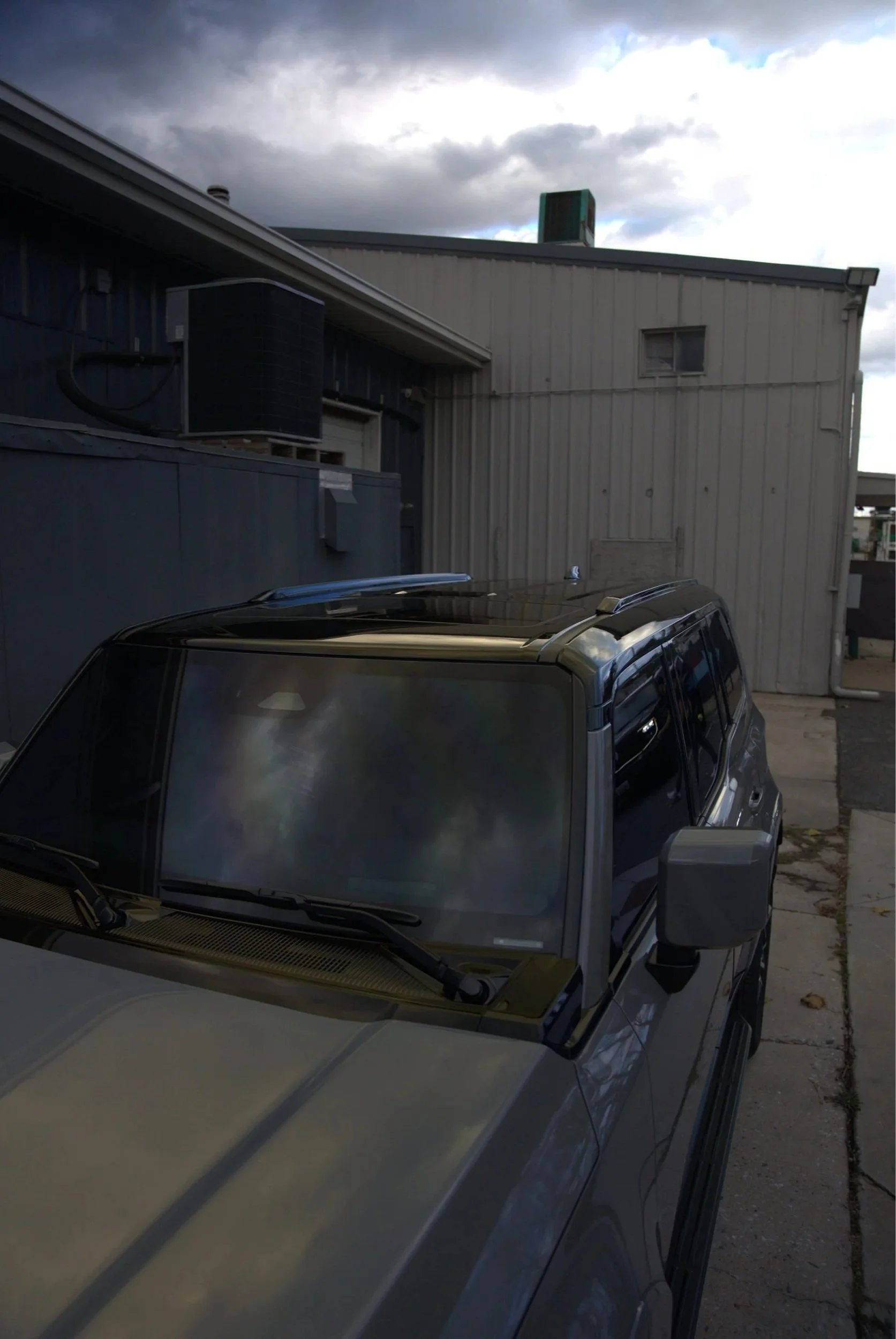 Part of a silver vehicle, possibly a SUV, parked outside a building with a beige metal exterior under a cloudy sky.