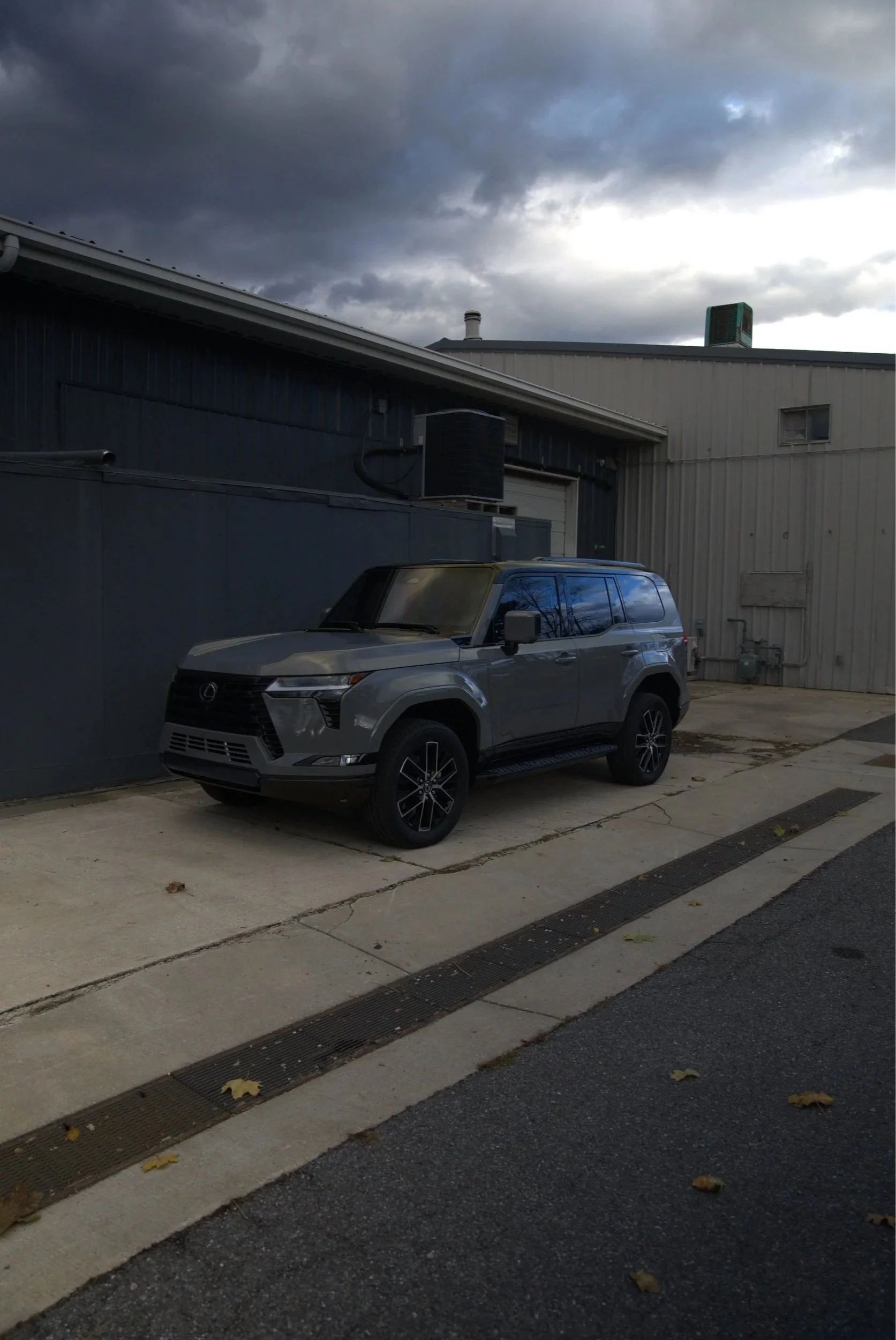 A gray Lexus SUV parked on a concrete sidewalk next to a dark blue industrial building under a cloudy sky.