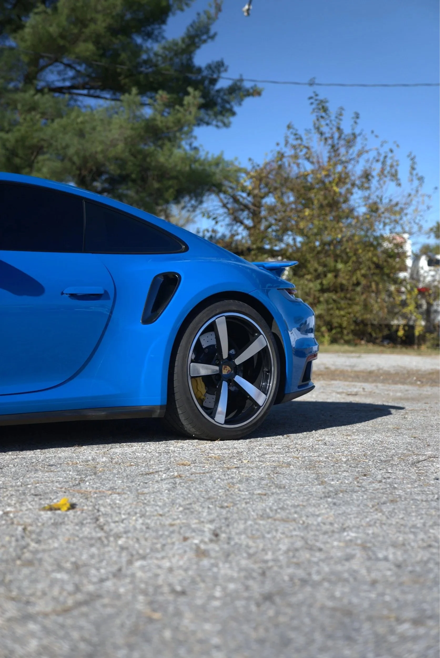 Close-up of a blue sports car parked on a gravel surface with trees and clear blue sky in the background.