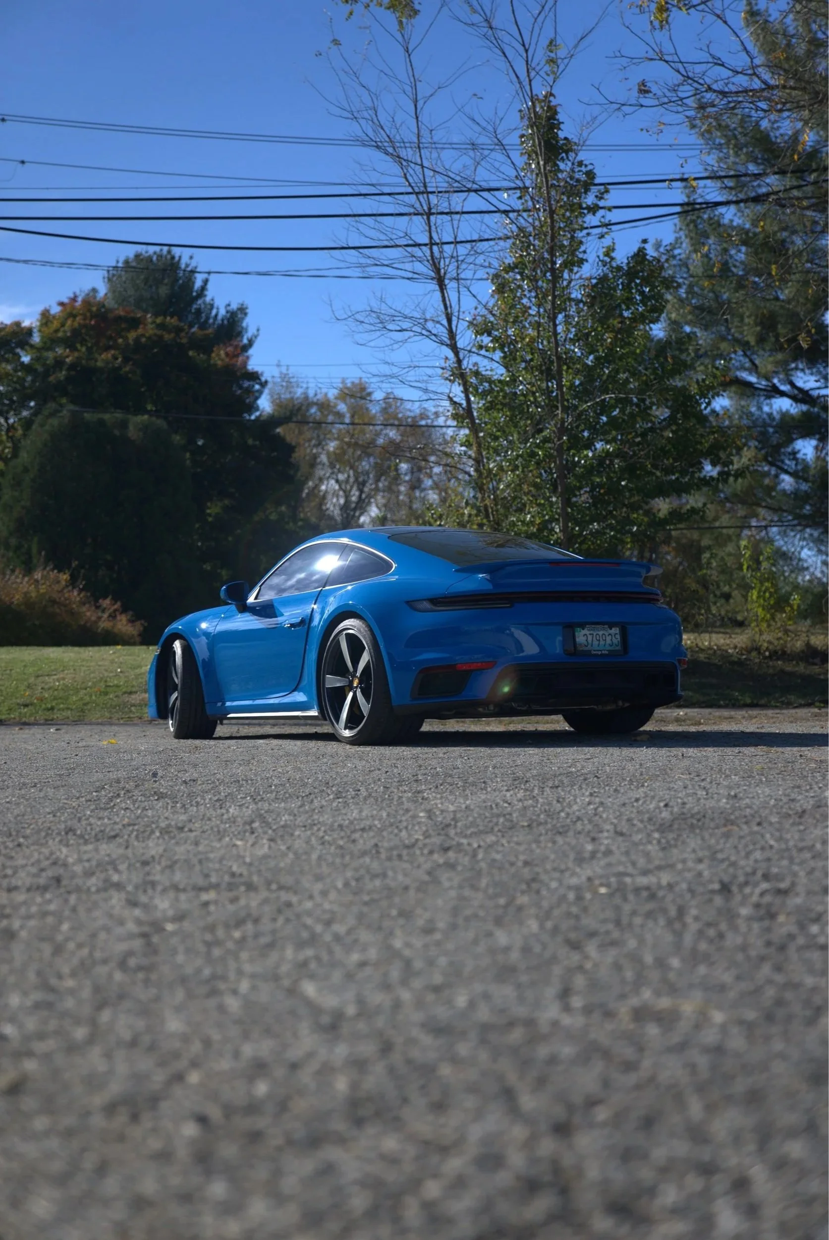Blue sports car parked on a gravel surface, with a background of trees and a clear blue sky.