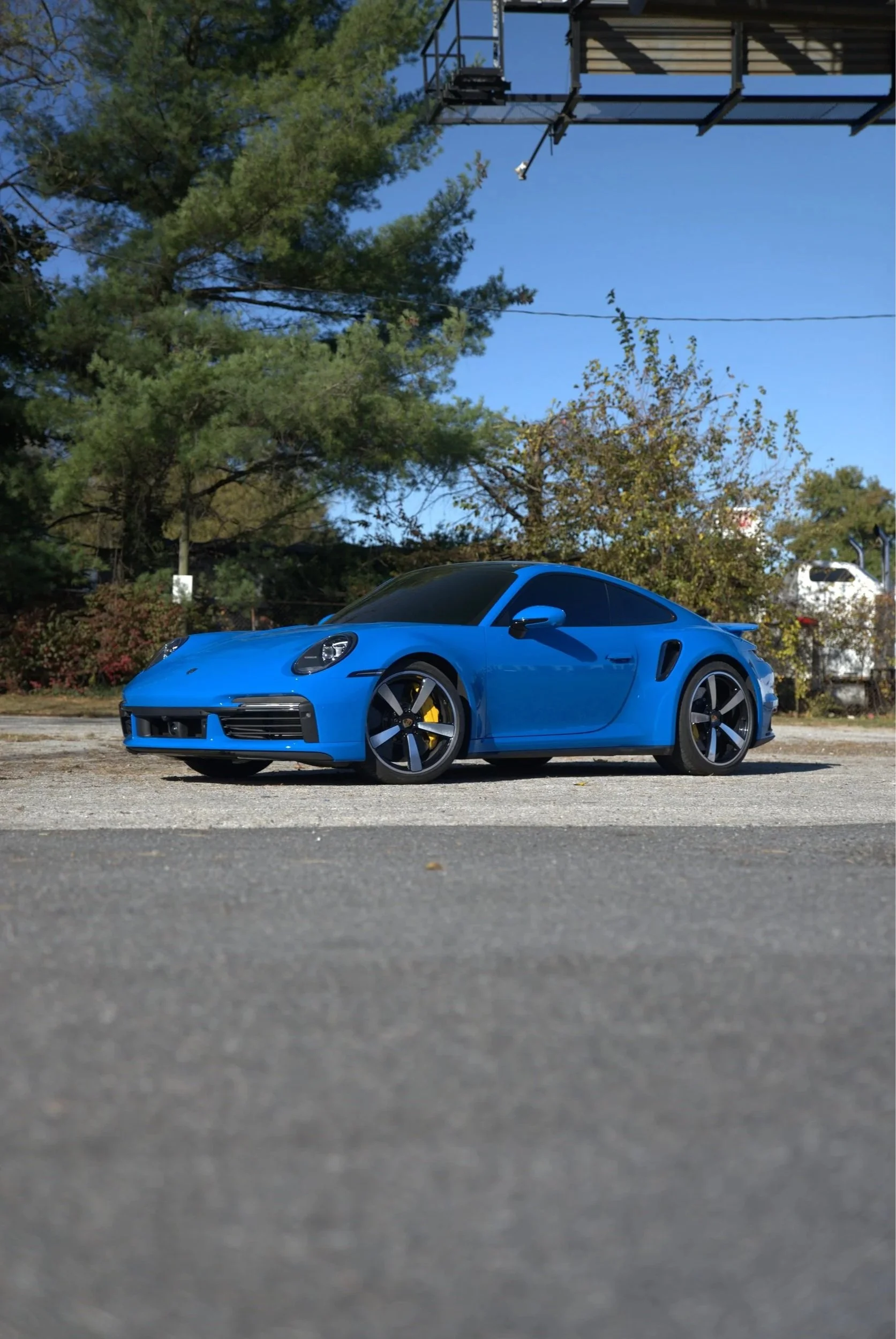 A blue sports car parked on an outdoor asphalt surface under a clear blue sky, with trees and a metal structure overhead.