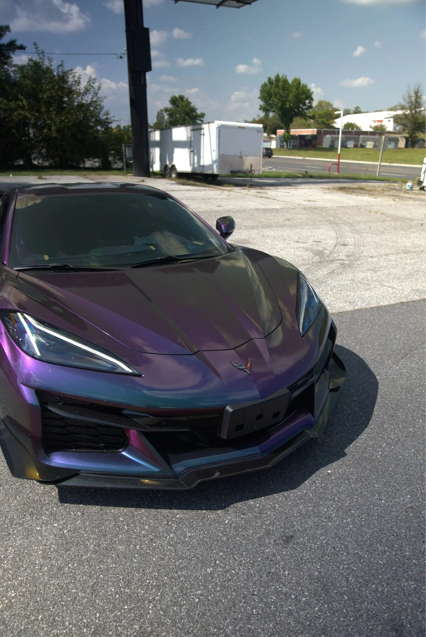 A purple sports car parked on a paved lot with a trailer in the background, trees, and a clear blue sky with scattered clouds.