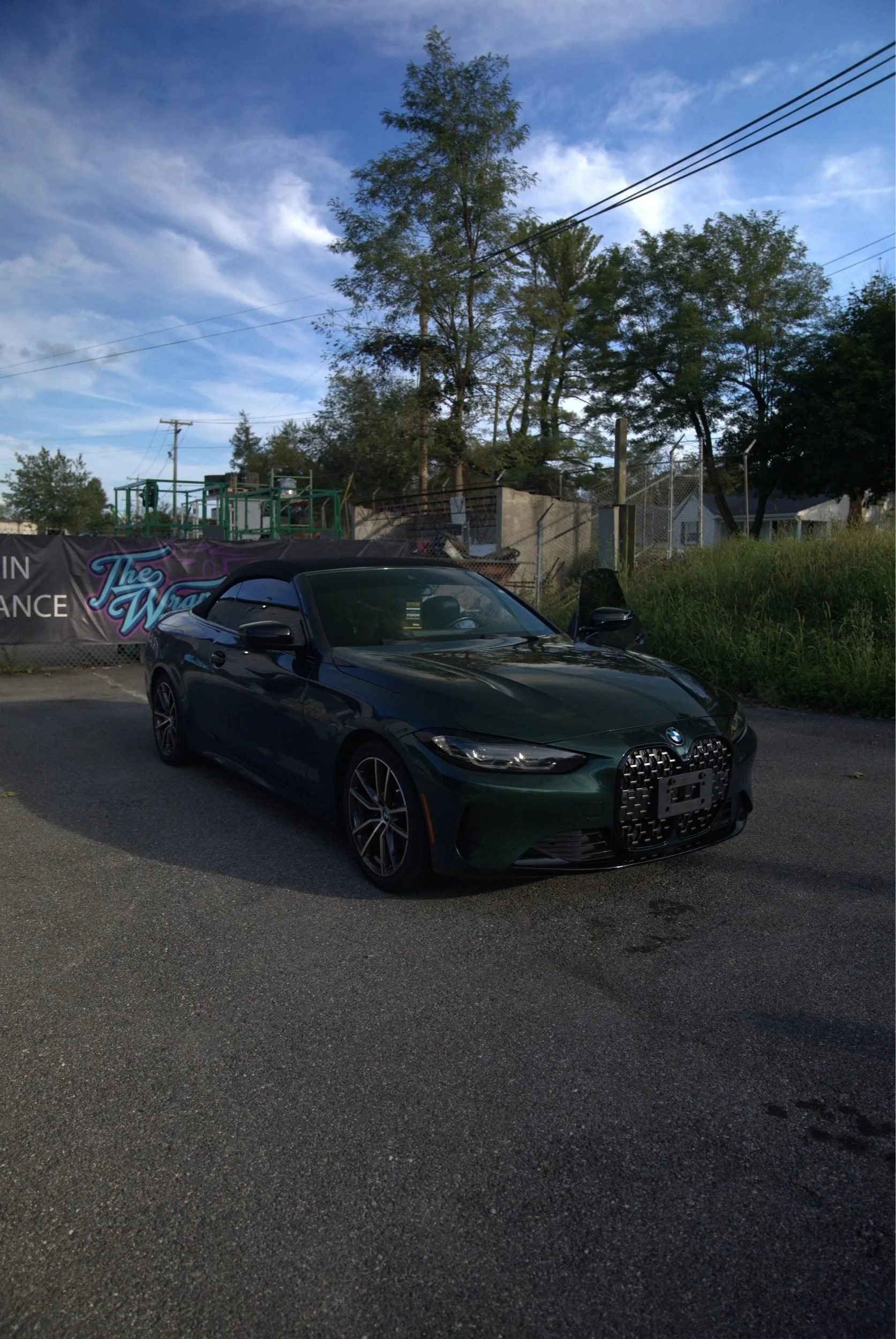 A dark green BMW convertible parked on a paved lot with a grassy hill, trees, and a fence in the background. The sky is partly cloudy.