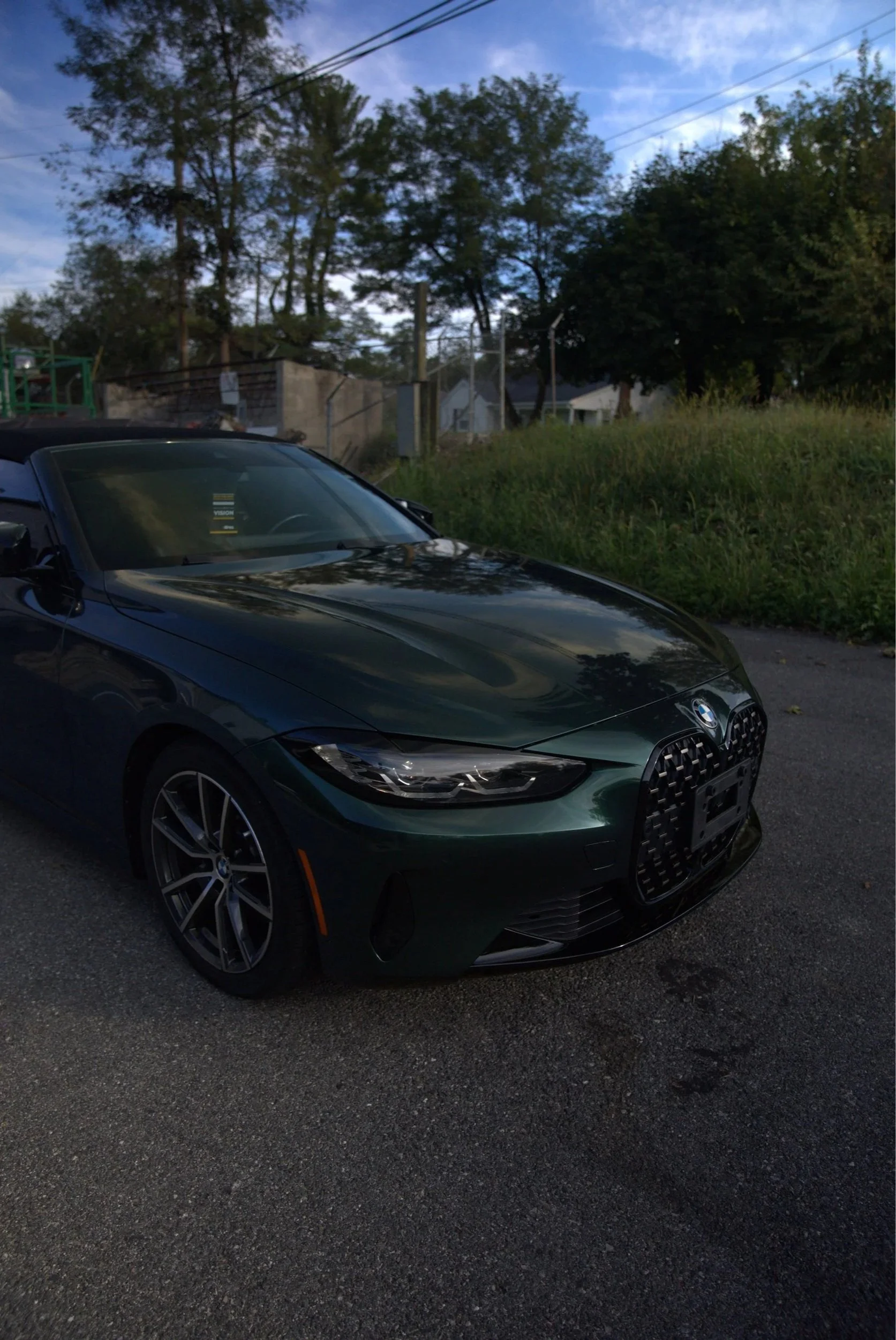 A dark green BMW convertible parked on the side of the road with a grassy area and trees in the background under a partly cloudy sky.