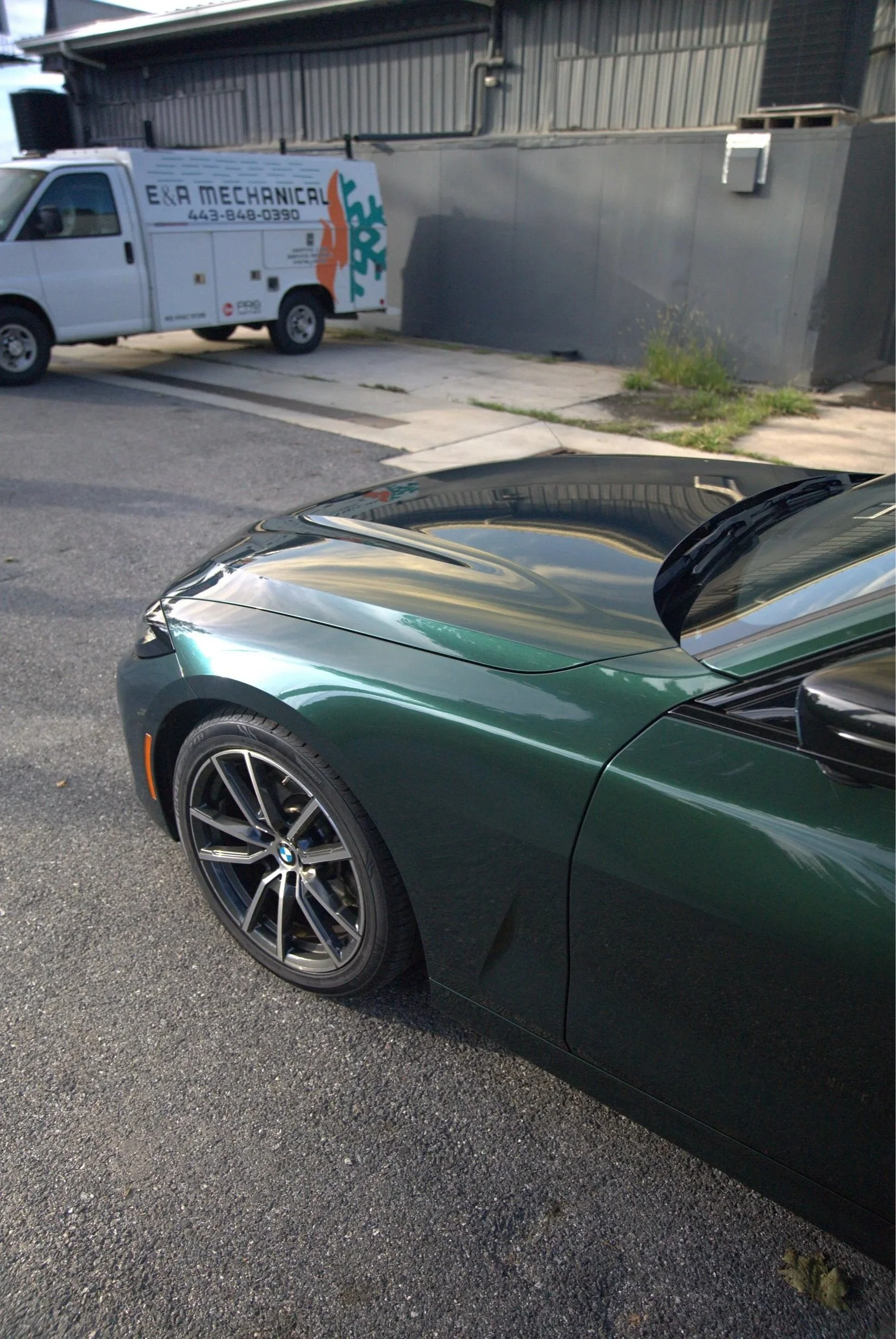Close-up of a green BMW sports car parked in front of a gray building with a white van in the background. The van has the text 'E&R Mechanical' and a phone number on its side.