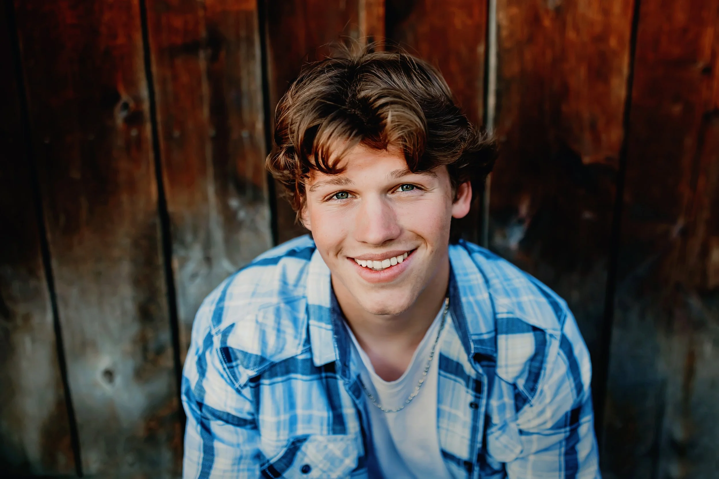 A high school senior smiles at the camera while wearing a blue plaid shirt over a white t-shirt, posed outdoors against a rustic wooden background in Bellingham, WA
