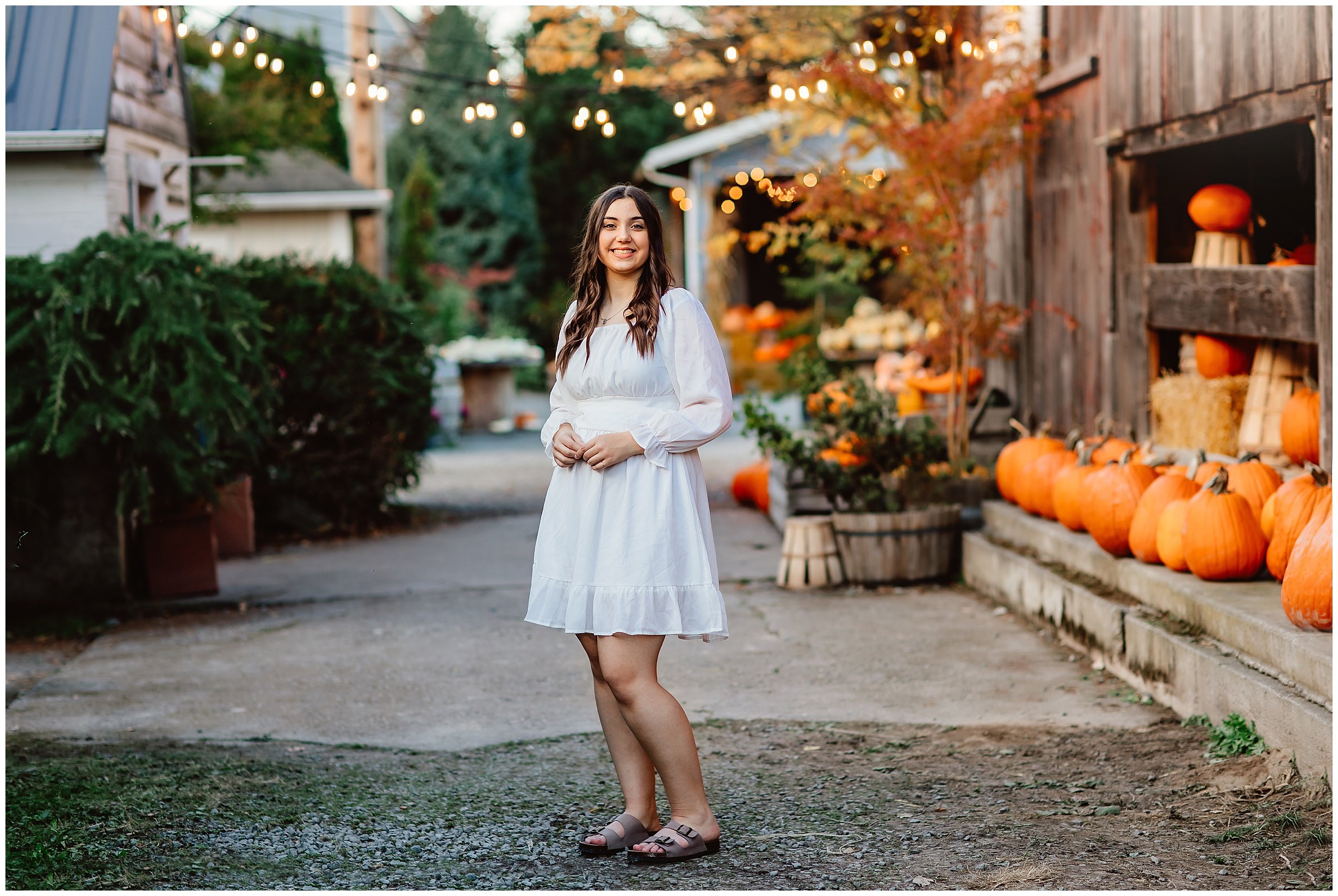  high school senior standing in a white dress smiling at the camera with string lights and pumpkins in the background 