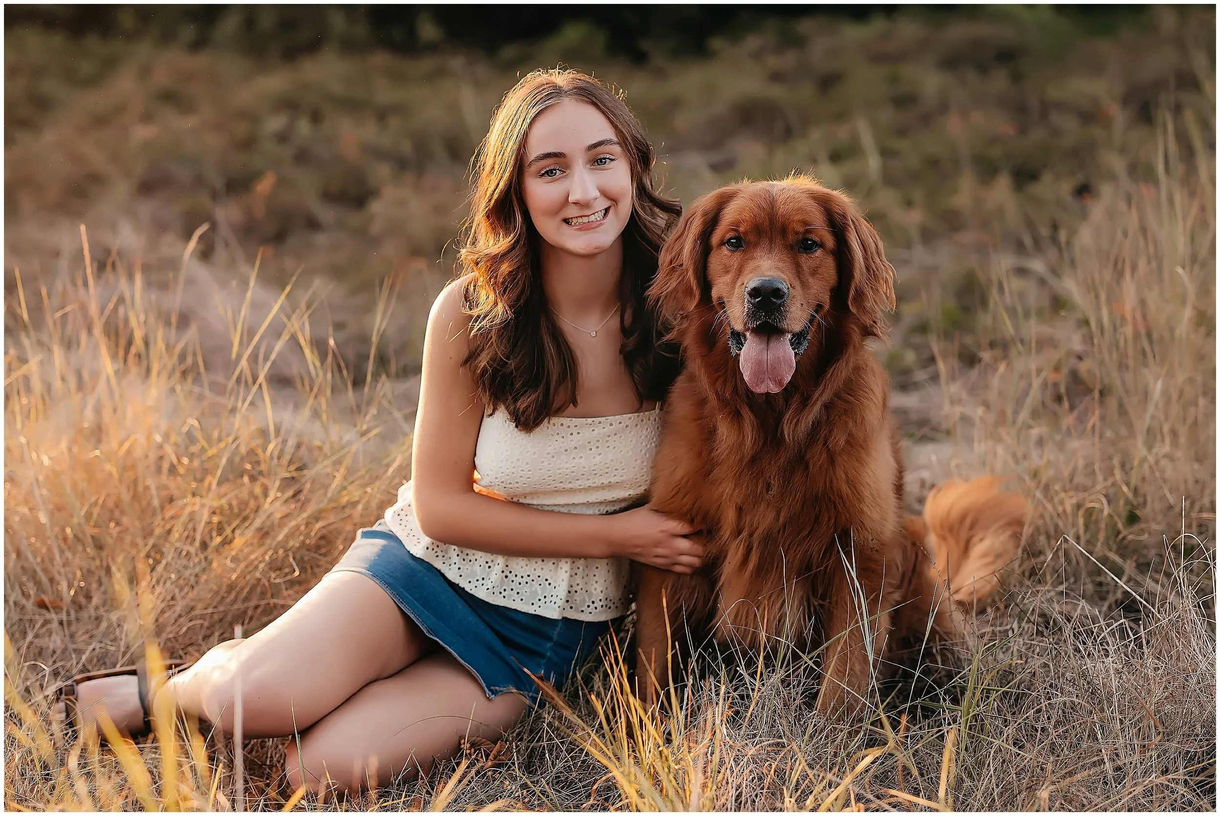 Senior portrait of a young woman sitting in a field with her golden retriever, softly lit by evening sunlight