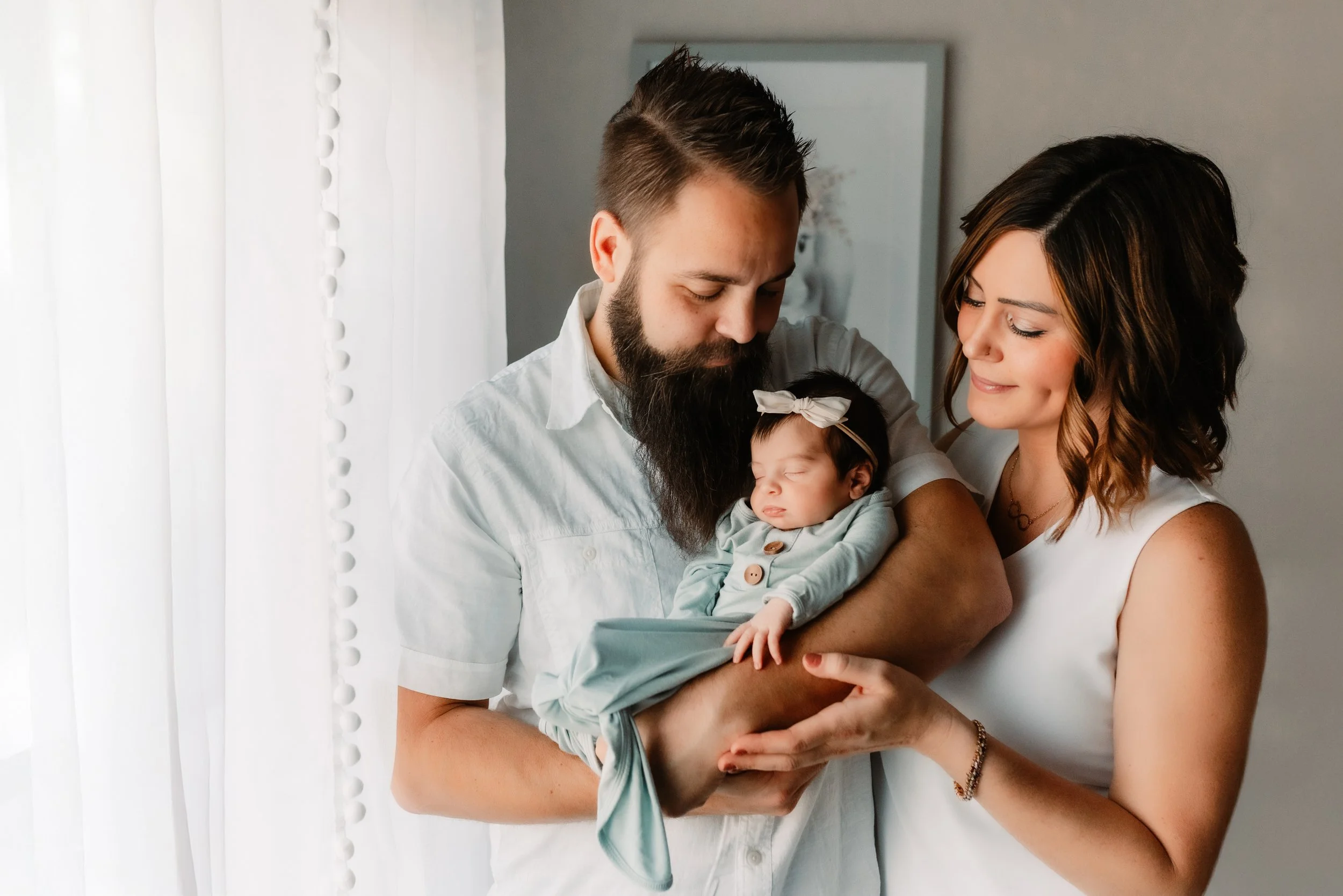 A mother and father gently hold their sleeping newborn baby indoors, looking down at her with soft smiles as she rests in her father's arms, wearing light green outfit and headband