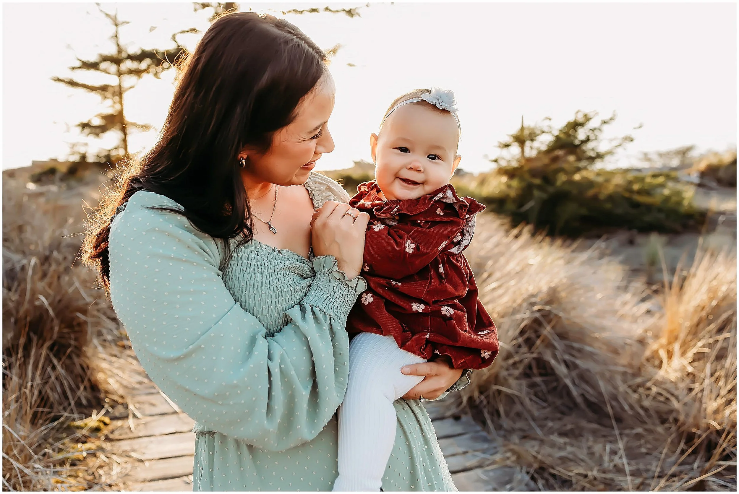 A mother holds her smiling baby daughter during an outdoor family photo session at Deception Pass State Park, standing on a sandy boardwalk with tall grass and trees in warm evening light