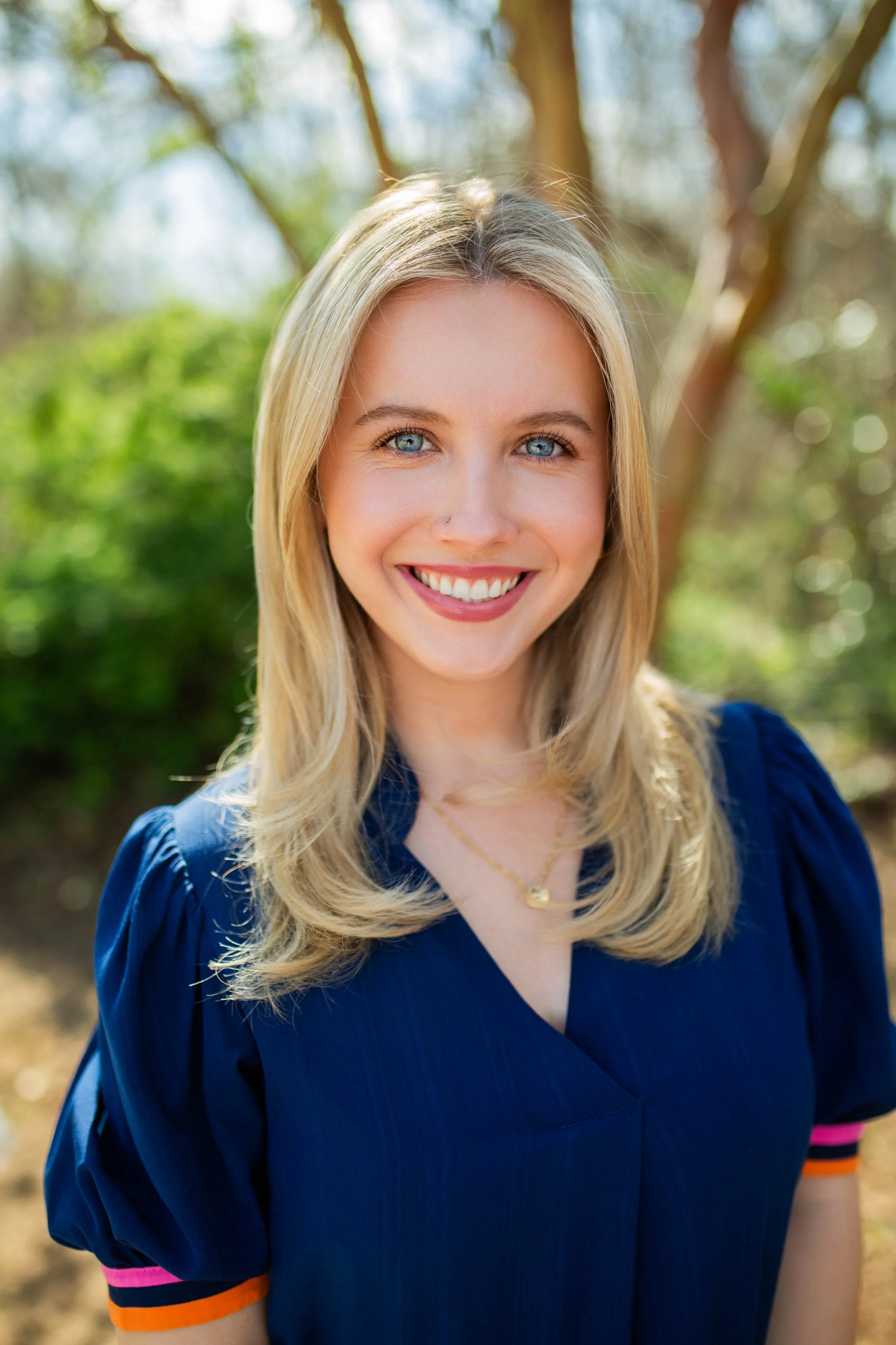 A young woman with blonde hair, blue eyes, and a nose piercing smiling outdoors on a sunny day.