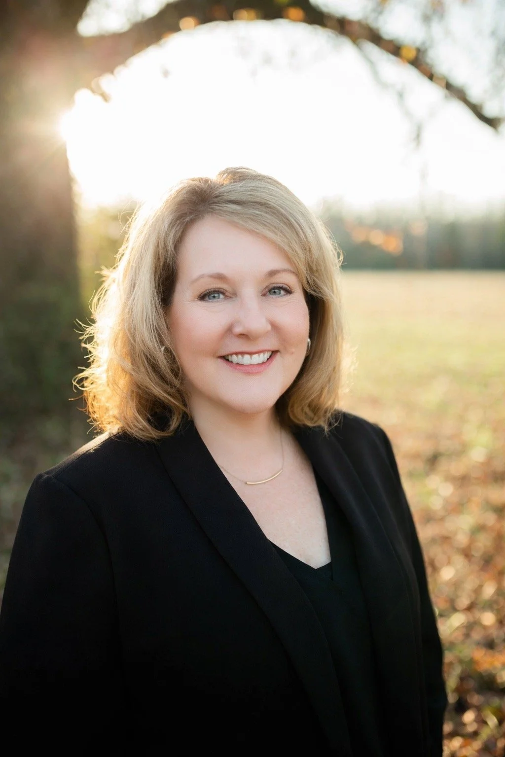 A female provisionally licensed social worker with shoulder-length blonde hair smiling outdoors in sunlight, wearing a black jacket and a delicate gold necklace, with a background of trees and a field.