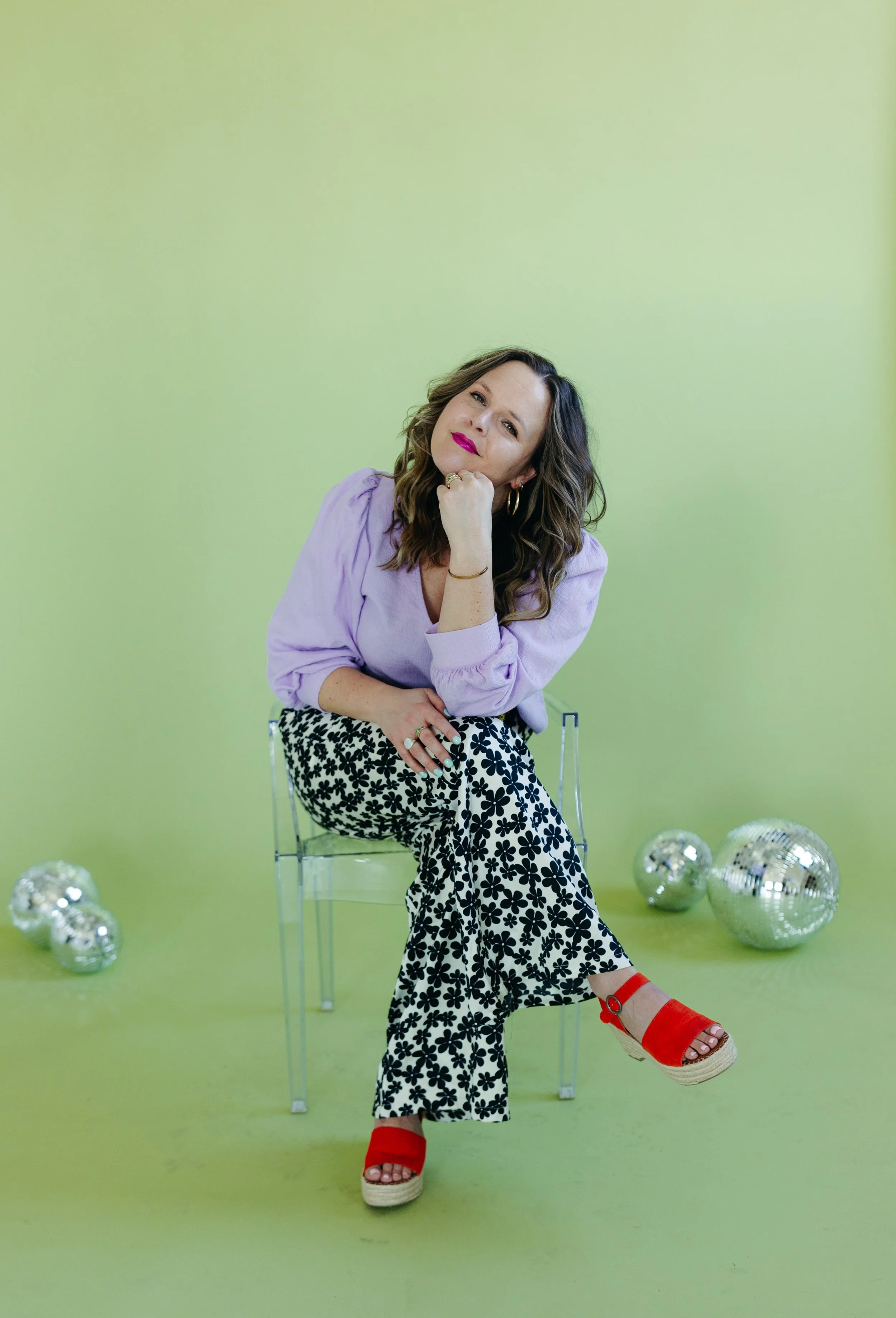 A woman with wavy brown hair and pink lipstick sits on a clear chair against a green background, wearing a light purple blouse, black and white floral patterned pants, and red platform sandals. She rests her chin on her right hand, looking at the camera, with silver disco balls on the floor around her.