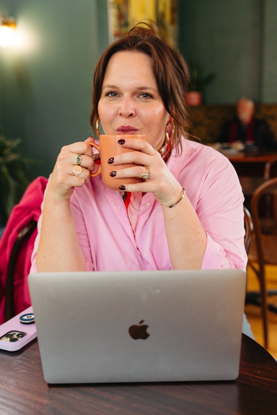 A woman with short brown hair and hoop earrings is sitting at a table, holding an orange mug, and looking at the camera. She wears a pink shirt, jewelry, and has a laptop and a phone on the table. The background shows a cafe with other people.