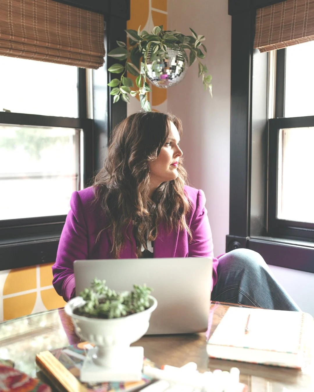 Woman with wavy brown hair and purple blazer sitting near a window, looking outside with a laptop and notebook on the table, decorated with a white plant pot and a disco ball hanging from the ceiling.