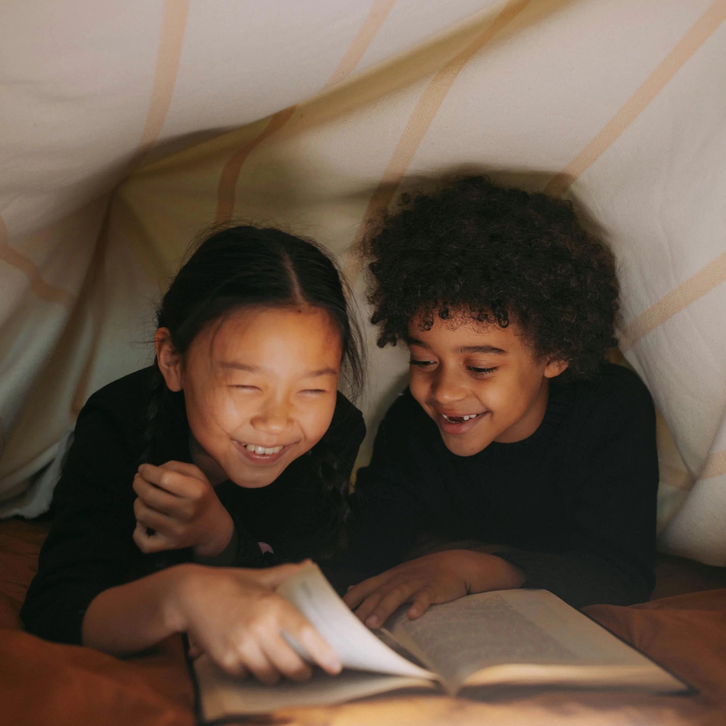 Two children, a girl and a boy, inside a blanket fort, reading a book and laughing.