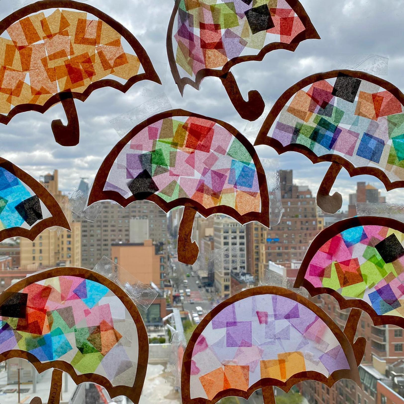 Colorful paper umbrellas with geometric patterns taped to a window, with a cityscape and cloudy sky in the background.