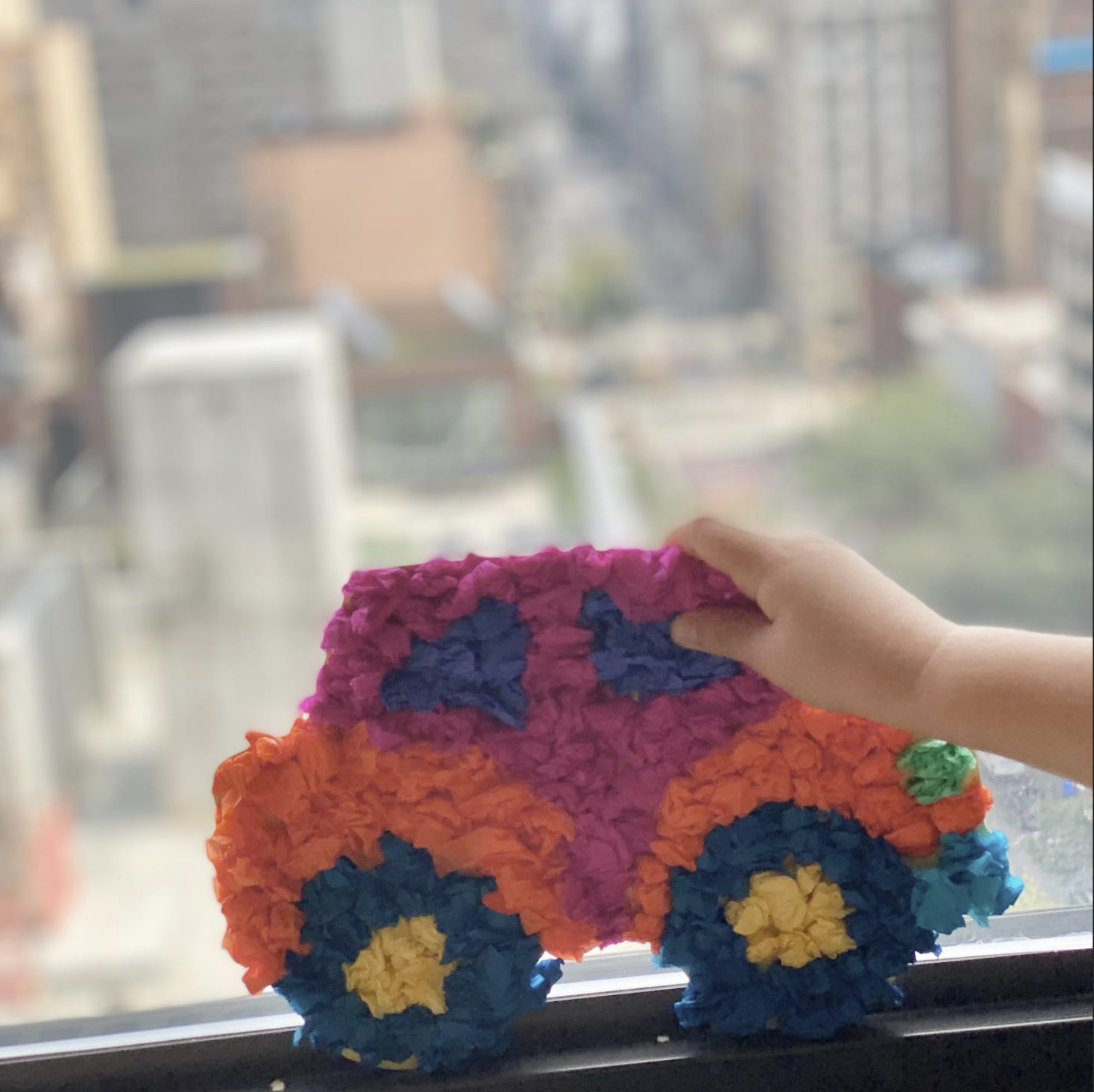 Colorful paper craft in the shape of a bus, held by a person's hand with a cityscape in the background.