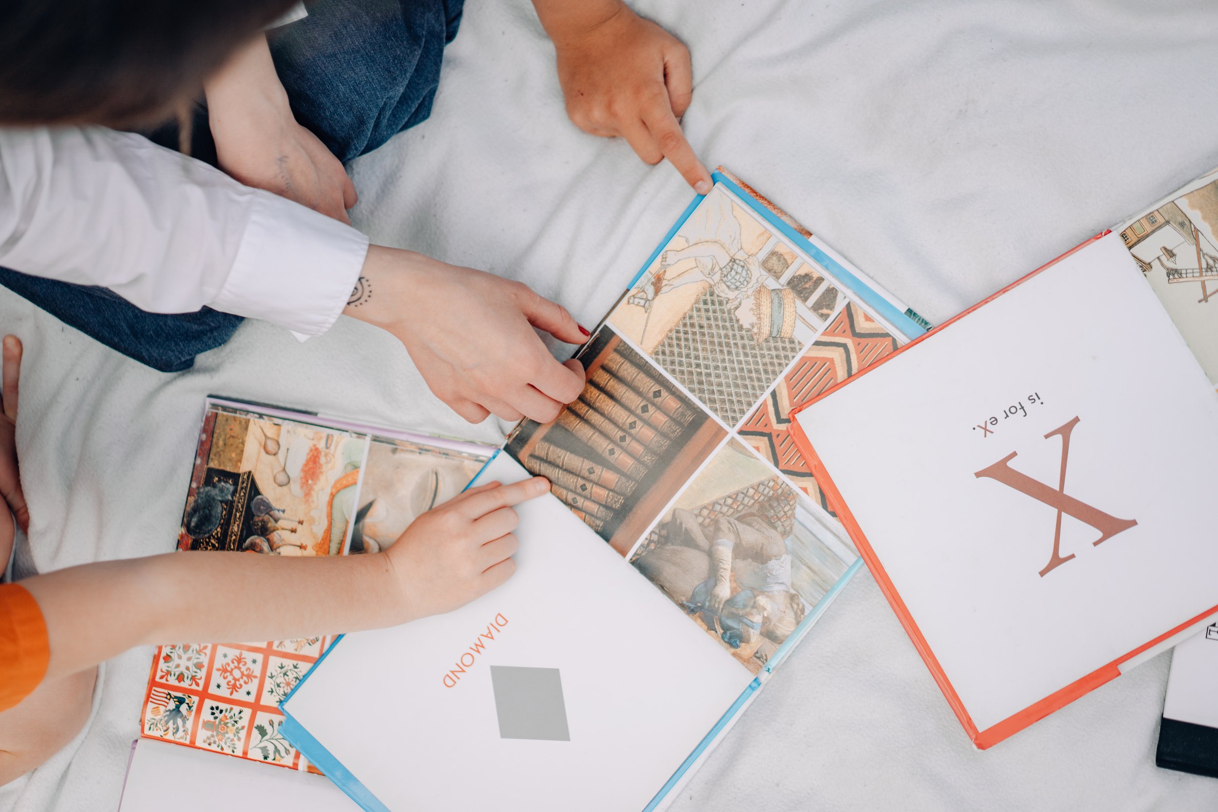 Two children looking at a photo book with colorful images, one child's hand pointing to a picture of a library. The book is on a white surface with other open books nearby.