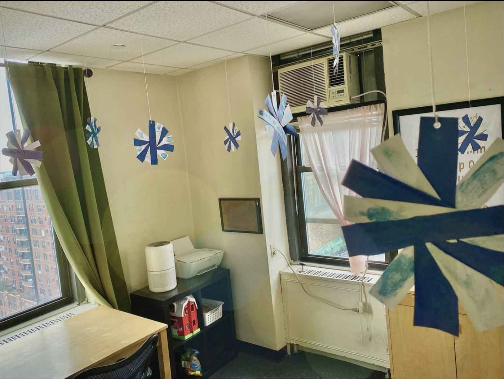 Decorative paper ornaments hanging from the ceiling in a room with beige walls, green curtains, and windows. A black shelf with a white humidifier, a red toy house, and other items is under the window. An air conditioning unit is mounted in the window, and a pink curtain covers part of the window.