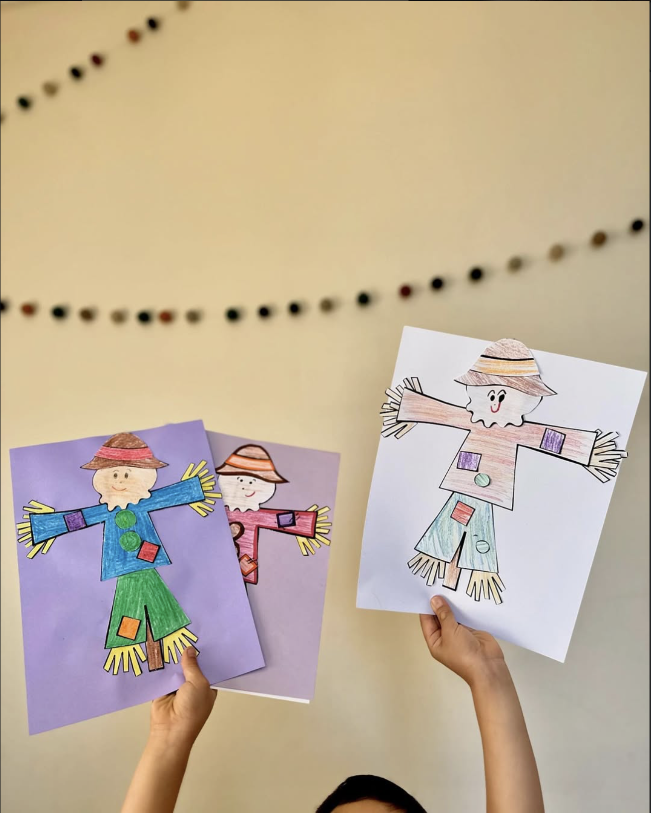 Three colored paper drawings of scarecrows being held up by children, with one drawing on white paper and two on colored paper backgrounds, against a beige wall with a string of colored decorations in the upper part.