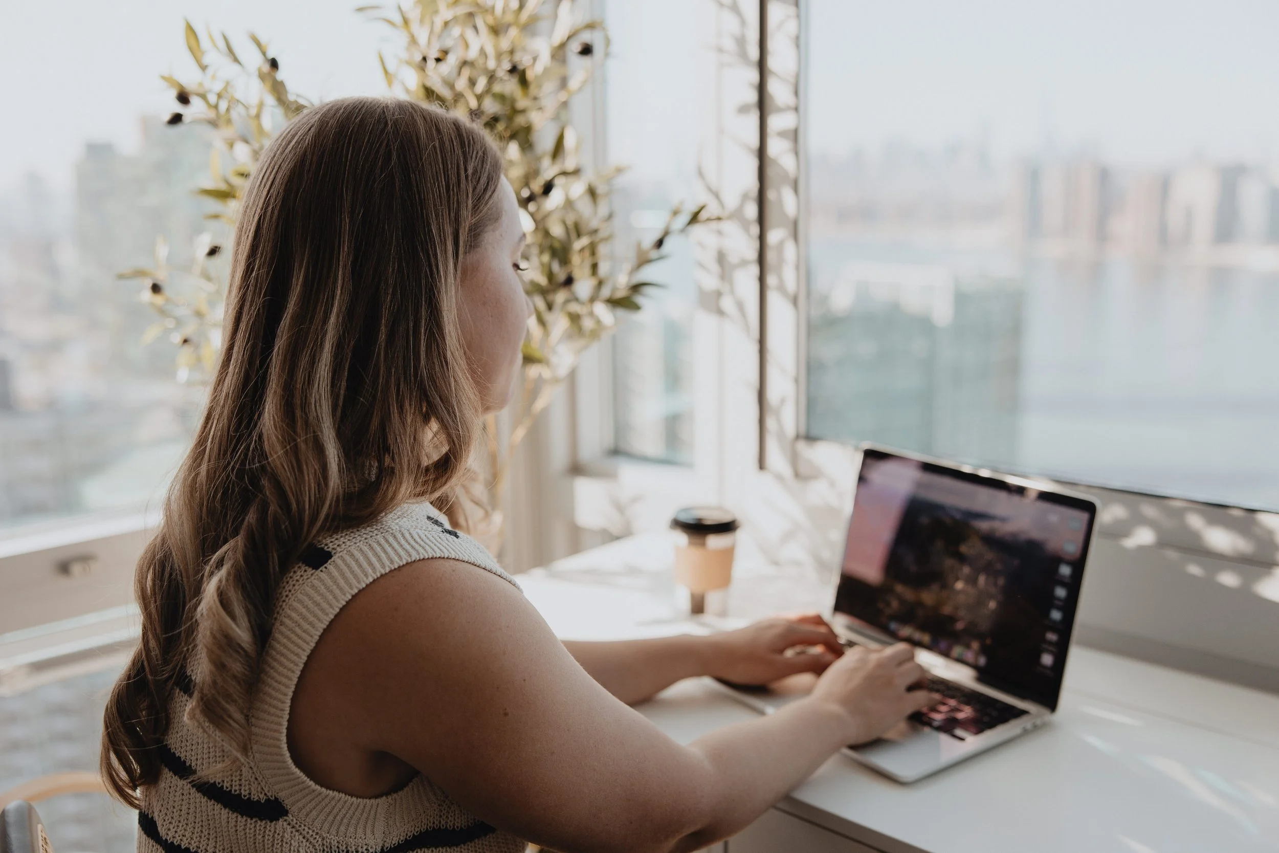 A woman with long brown hair working on a laptop at a white desk near a large window with a city view, a takeaway coffee cup beside her.