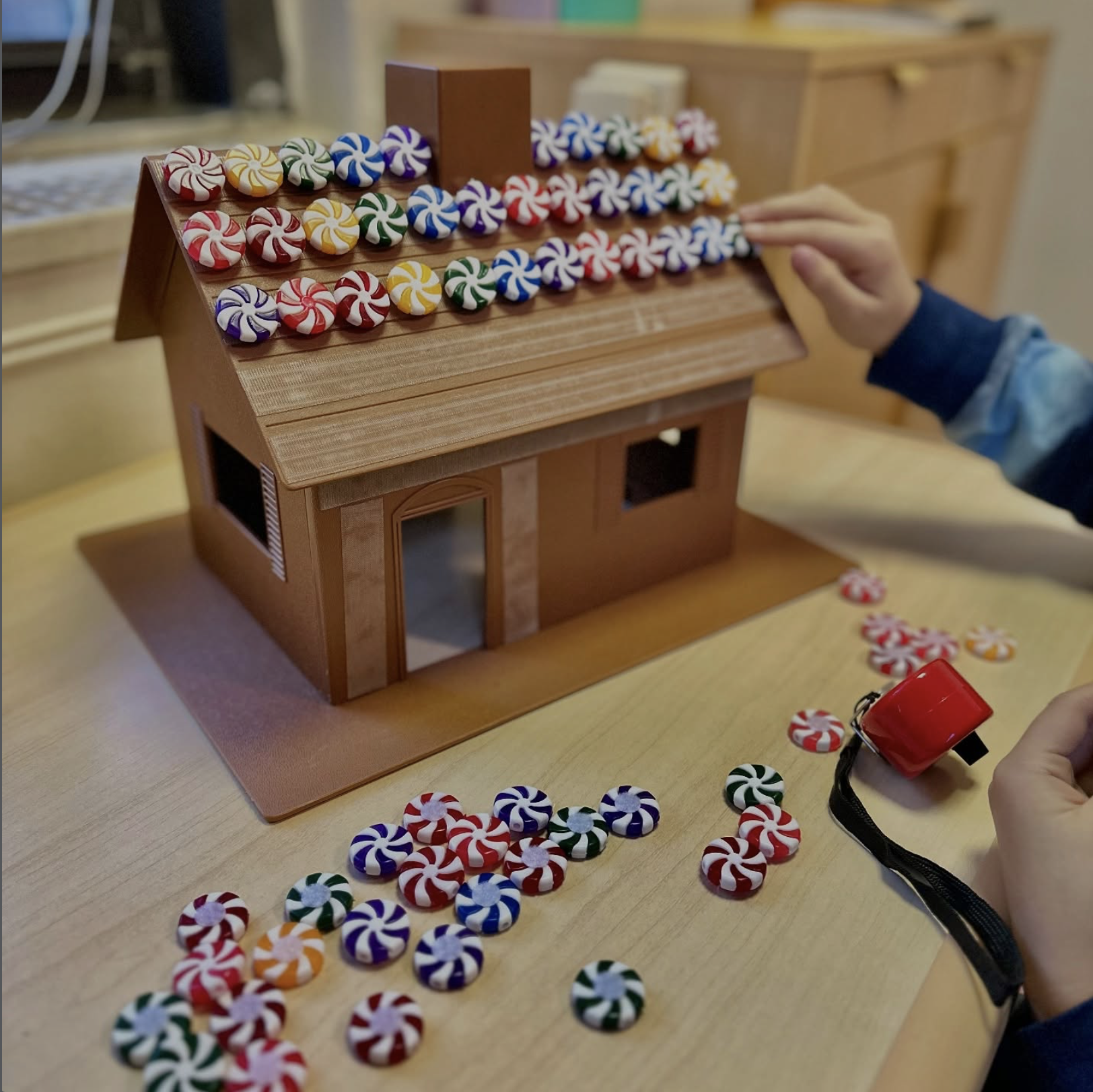 A small wooden house-shaped tabletop marble run with peppermint candy-shaped marbles in red, white, green, purple, and orange colors. A person's hand adjusts the top row of marbles, and another hand with a red marble launcher is visible near the bottom.