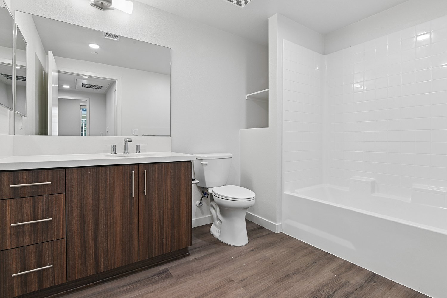 Bathroom in Rendezvous Phase II apartment home with wood-style plank flooring, large vanity with dark wood cabinetry and quartz countertops, a shower/tub, and a storage nook.