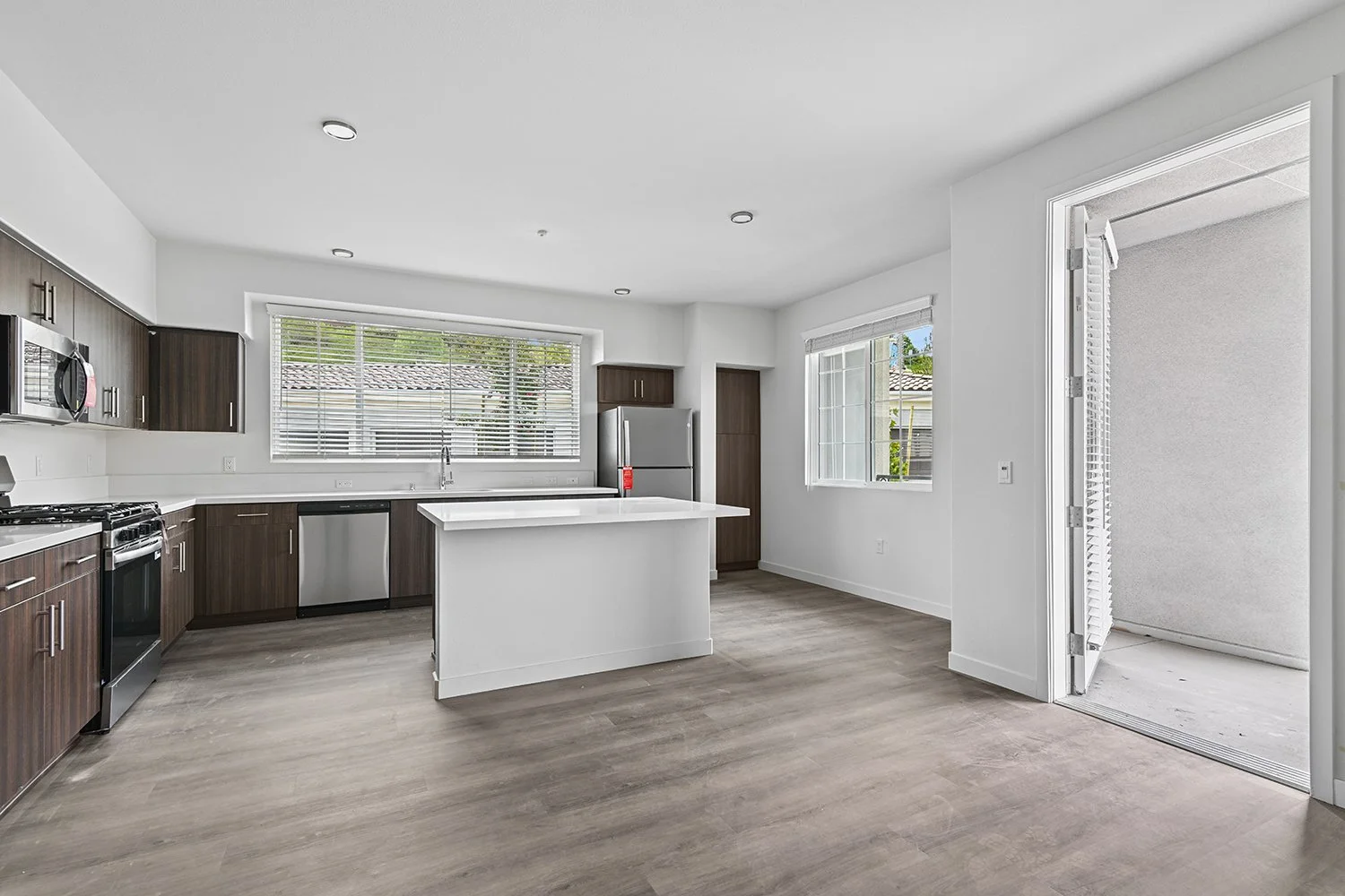 Spacious open concept living area leading to kitchen in Rendezvous Phase II apartment home. Wood-style plank flooring, modern dark walnut cabinetry, stainless steel appliances, quartz countertops, island, and a lot of windows allowing for gorgeous na