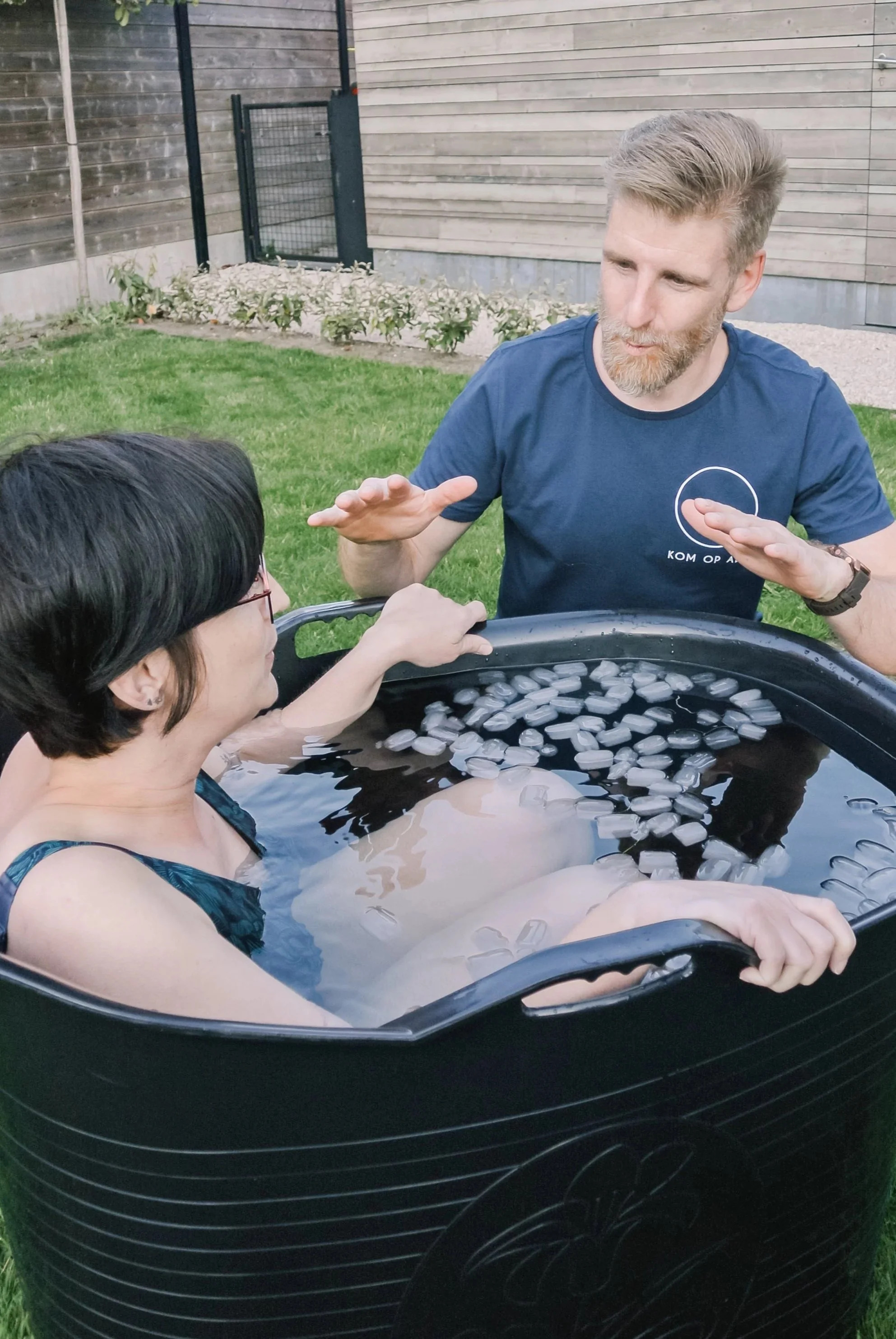 A woman with short black hair and glasses sitting in a large black container filled with water and ice cubes, outdoors in a backyard. A man standing beside the container is gesturing while speaking to her.