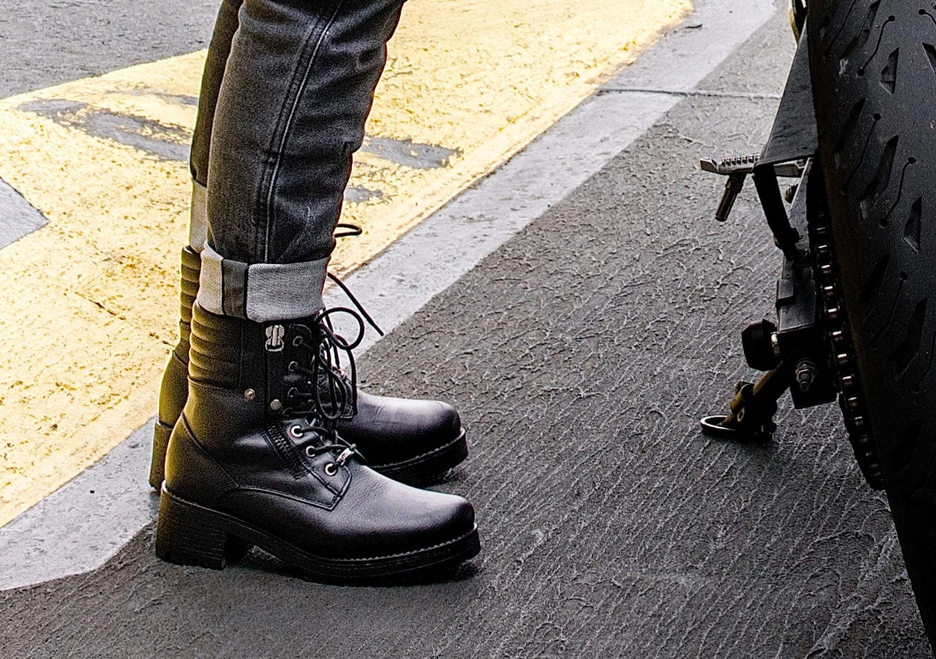 Close-up of a person wearing black combat boots and black jeans standing next to a motorcycle on a paved surface.