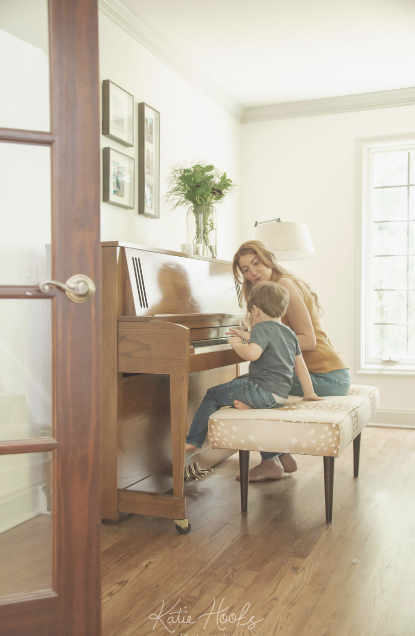 A woman and a young boy sitting together on a padded bench in front of a wooden piano in a brightly lit room with a large window.