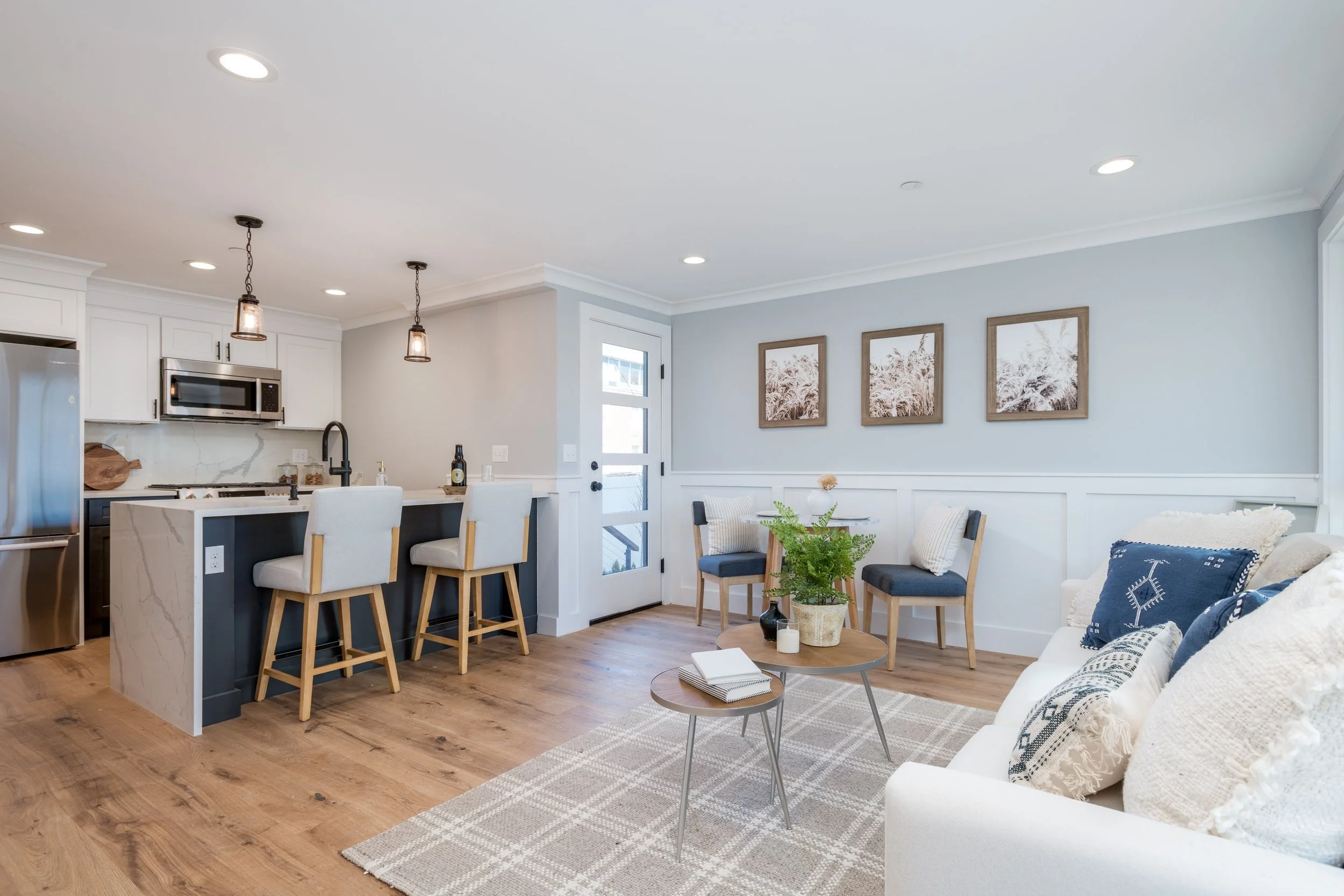 Open concept living room and kitchen with white cabinetry, stainless steel appliances, a marble island with bar stools, a gray wall with framed artwork, a white sofa with patterned pillows, and a small round table with plants and books.