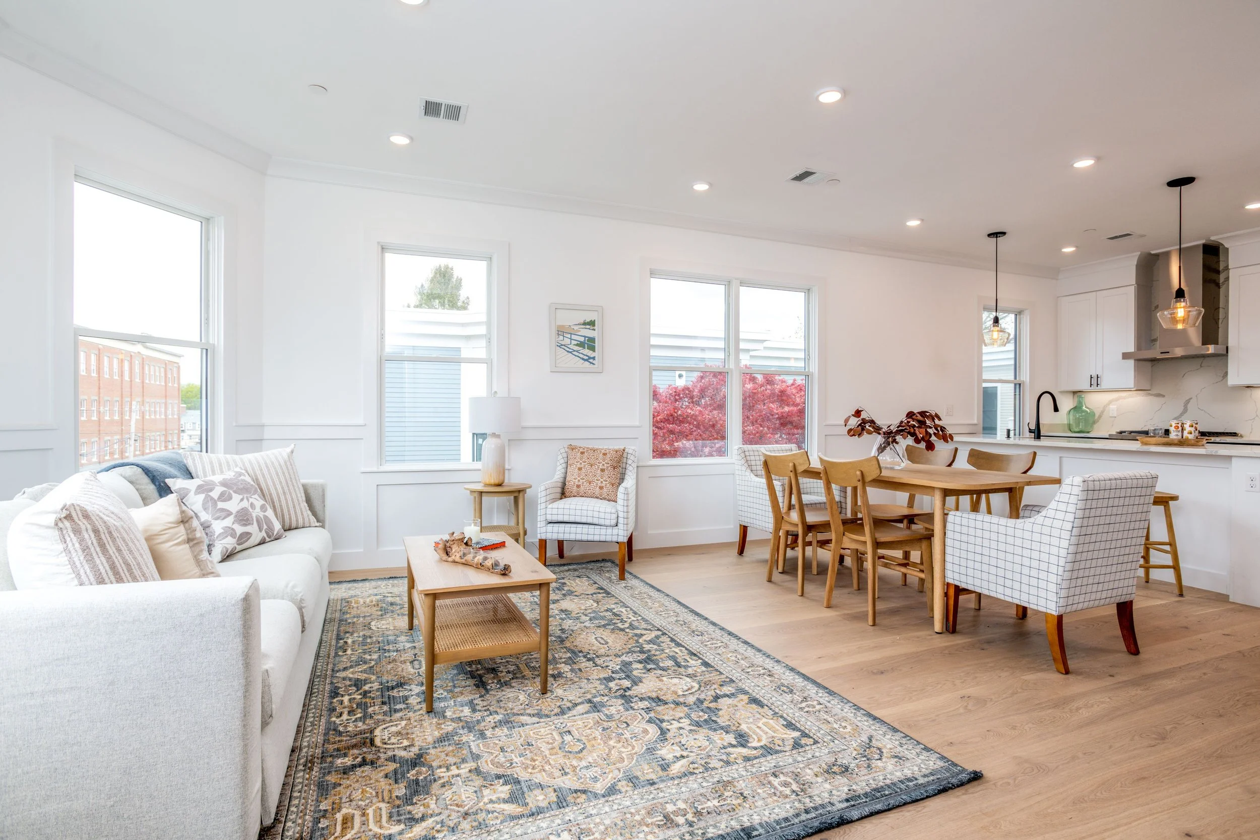 Open-concept living and dining area with white walls, hardwood flooring, large windows, beige sofa with pillows, wooden coffee table, patterned area rug, armchair, with dining table, chairs, and kitchen counters in the background.