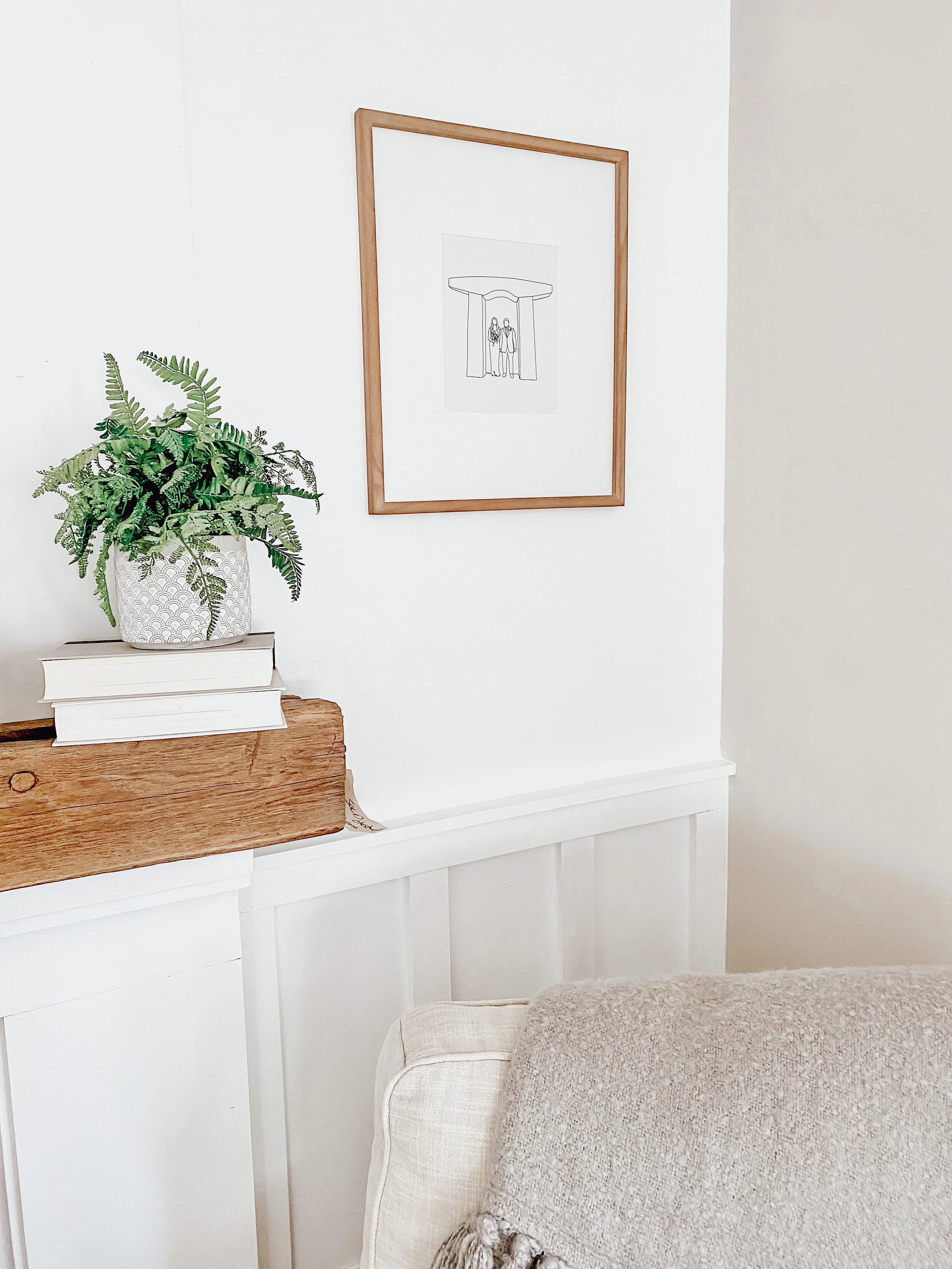 Interior of a living room corner with a wooden console table holding a potted fern plant and two stacked books, framed minimalist line drawing on the white wall, beige sofa with a textured throw blanket.